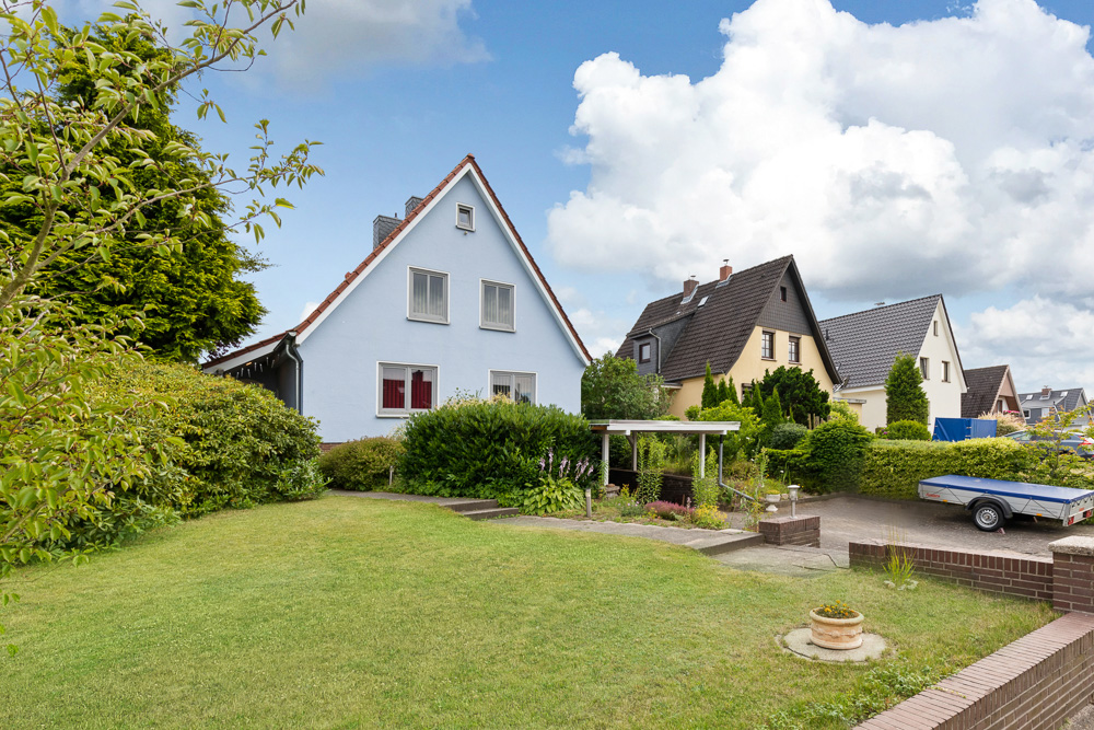 Zwei Einfamilienhäuser mit gepflegtem Garten und Vorgarten, daneben eine gepflasterte Einfahrt mit Anhänger.