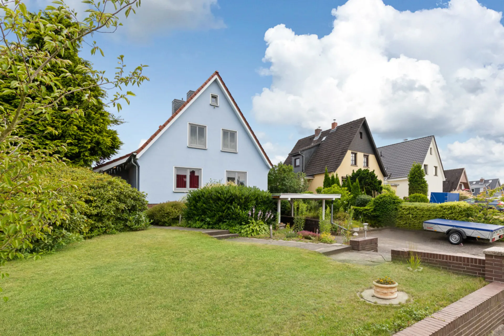 Zwei Einfamilienhäuser mit gepflegtem Garten und Vorgarten, daneben eine gepflasterte Einfahrt mit Anhänger.