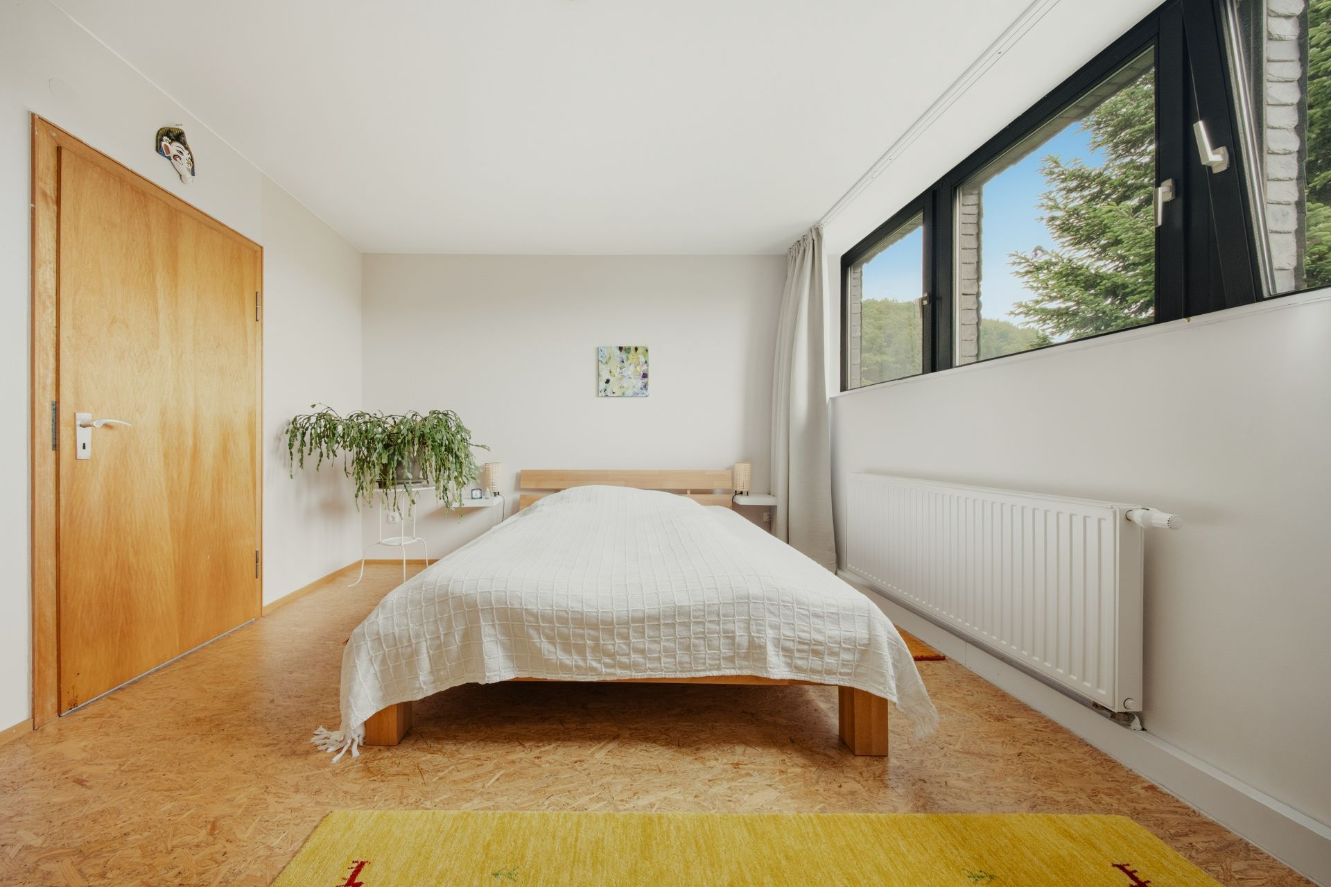 Helles Schlafzimmer mit Einzelbett, großem Fenster, Pflanze und Holzboden.