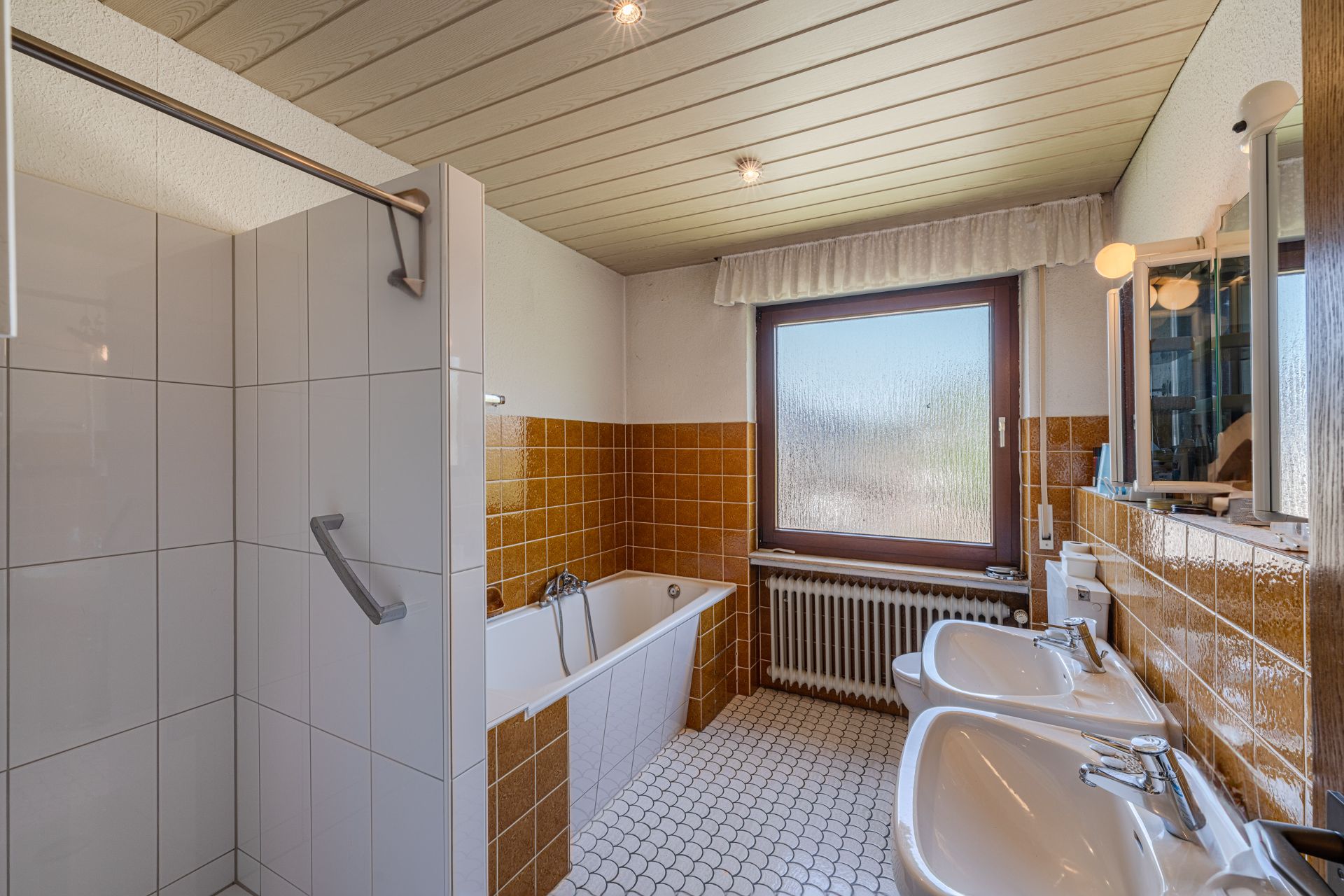 Badezimmer mit braunen Fliesen, Badewanne, separater Dusche, Doppelwaschbecken und großem Fenster.