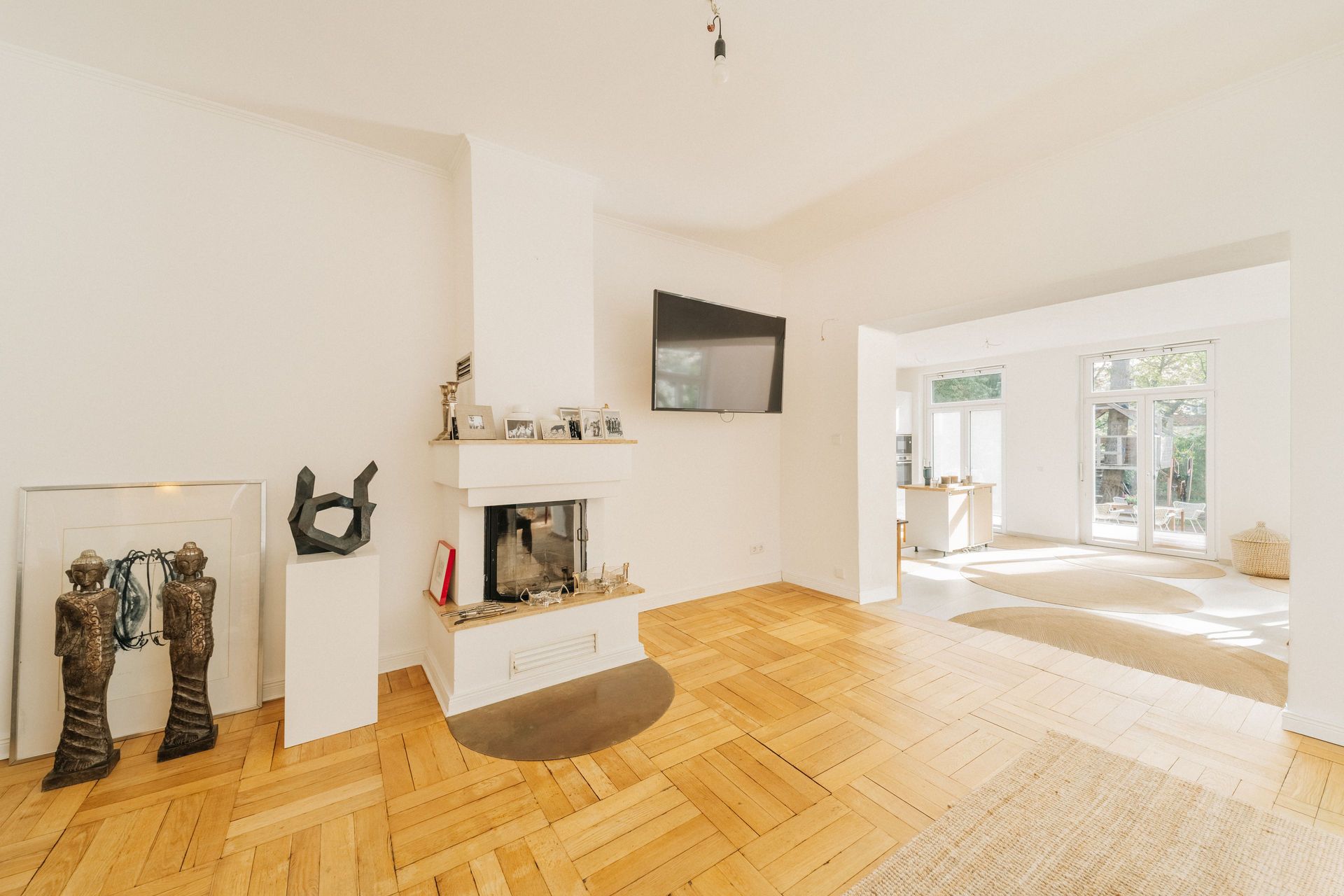 Helles Wohnzimmer mit Kamin, Parkettboden, Fernseher und großen Fenstern mit Blick in den Garten.