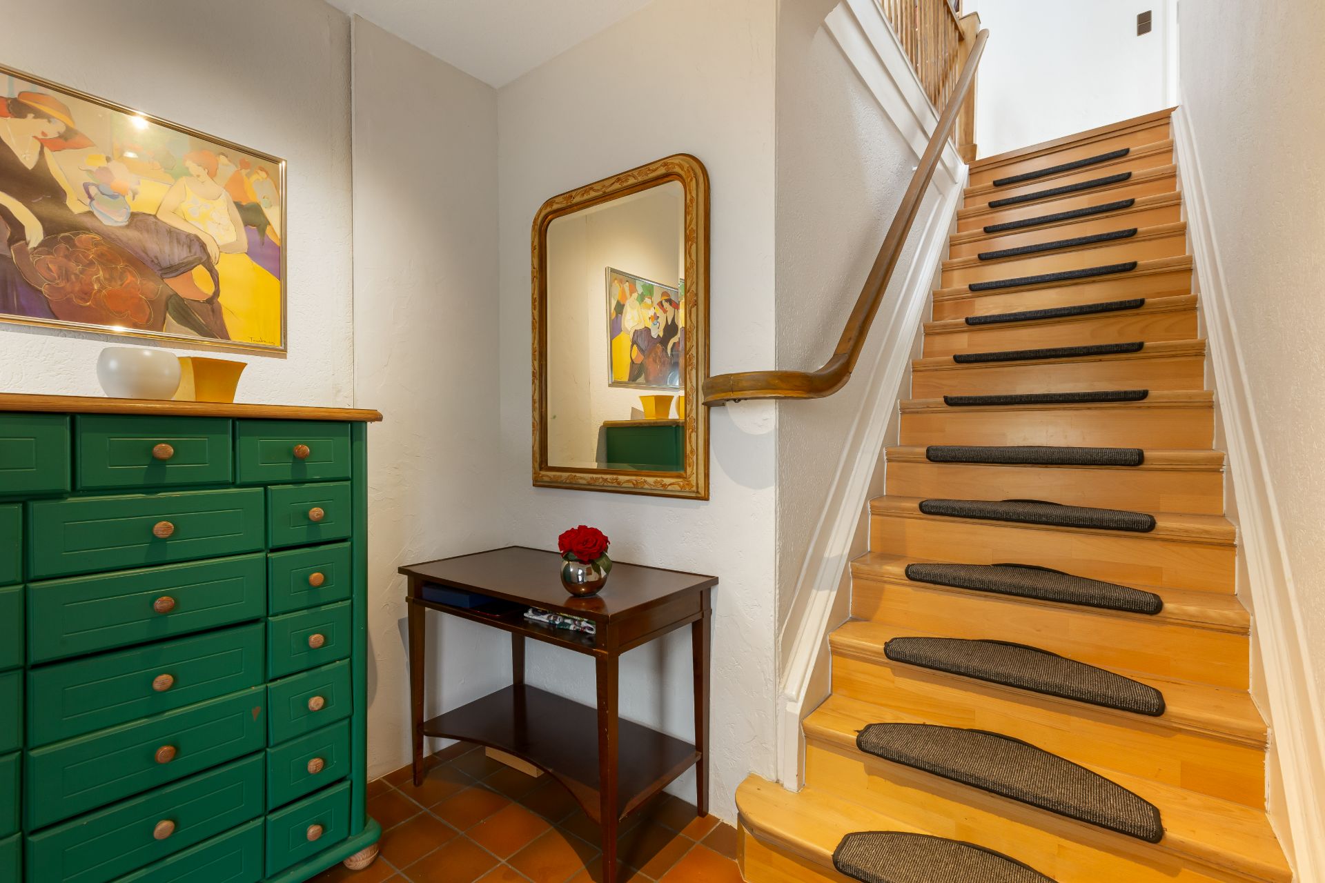 Hausflur mit Holztreppe, grünem Sideboard, Wandspiegel und kleinem Tisch mit Vase.
