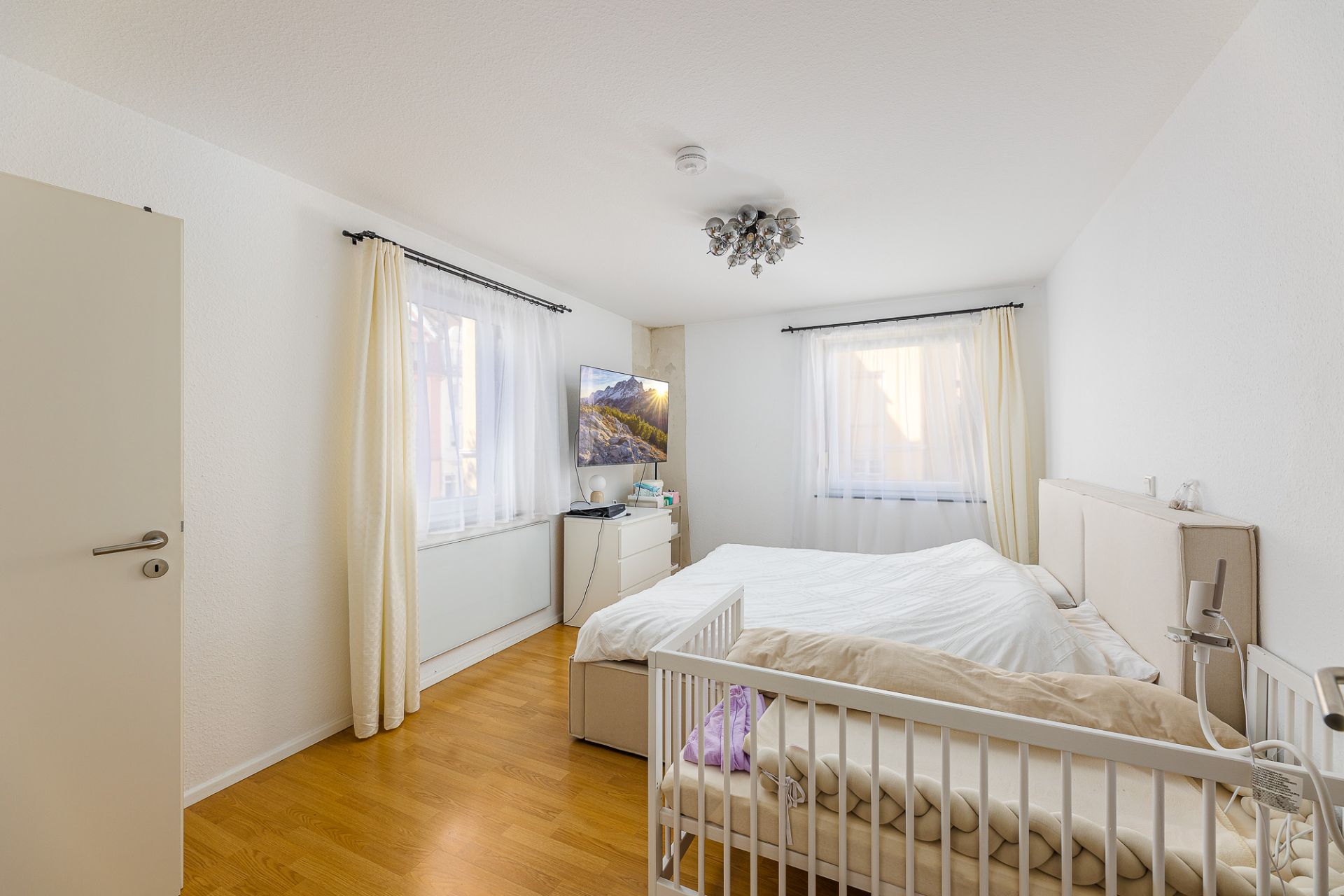 Helles Schlafzimmer mit Doppelbett, Babybett, zwei Fenstern und Holzboden.