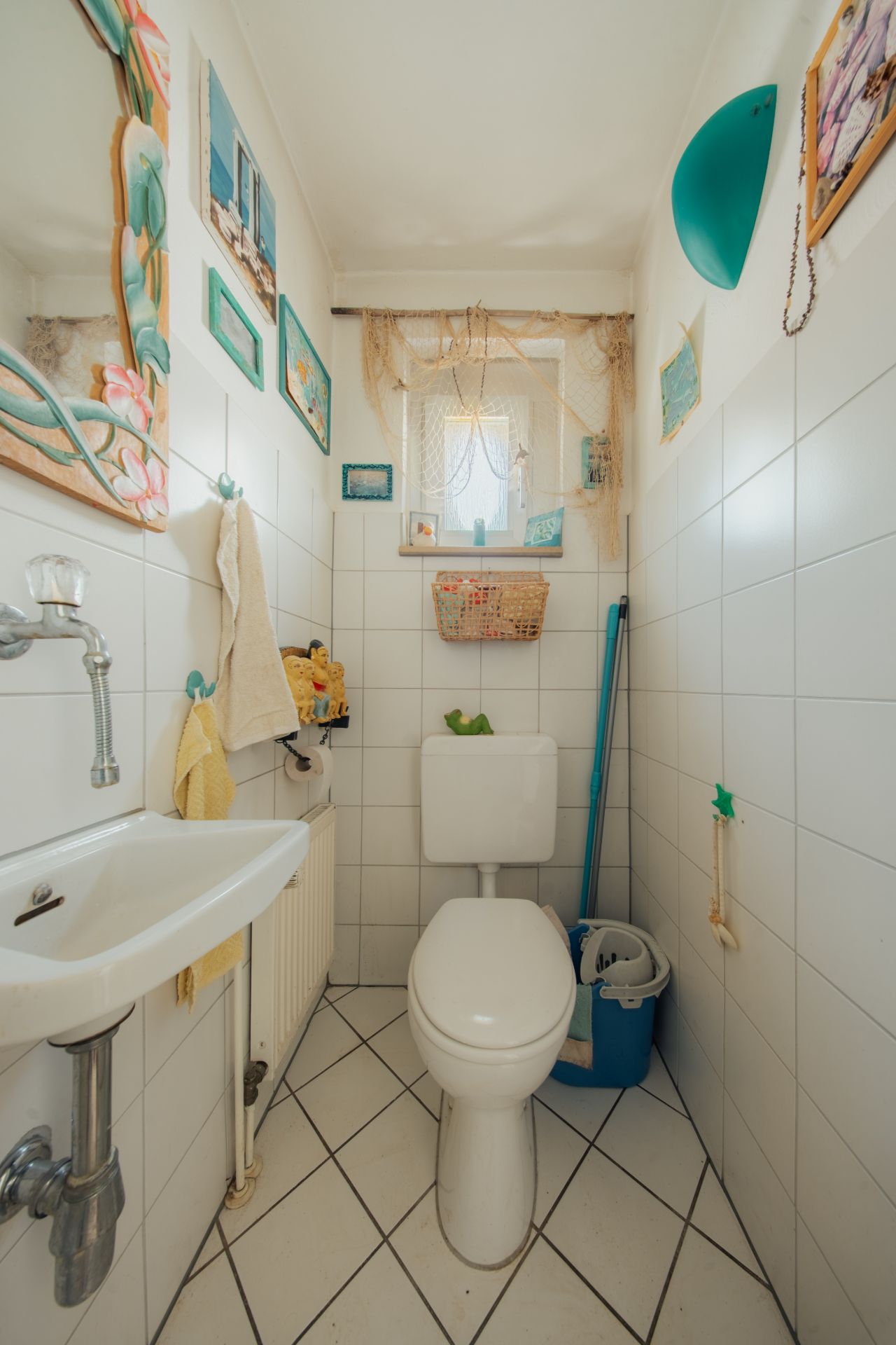 Kleines Badezimmer mit Toilette, Waschbecken, Wandbildern und Putzutensilien auf gefliestem Boden.