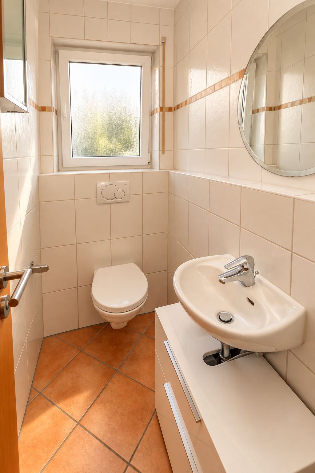 Kleines Badezimmer mit Wandtoilette, Waschbecken, rundem Spiegel und Fenster.