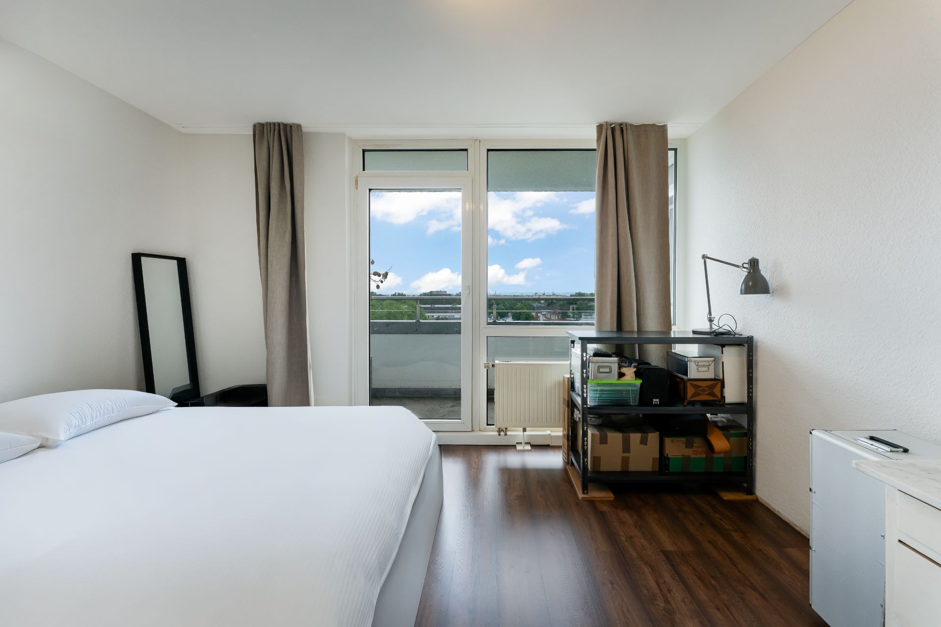 Helles Schlafzimmer mit Bett, Spiegel, Regal, Holzfußboden und Balkonzugang.