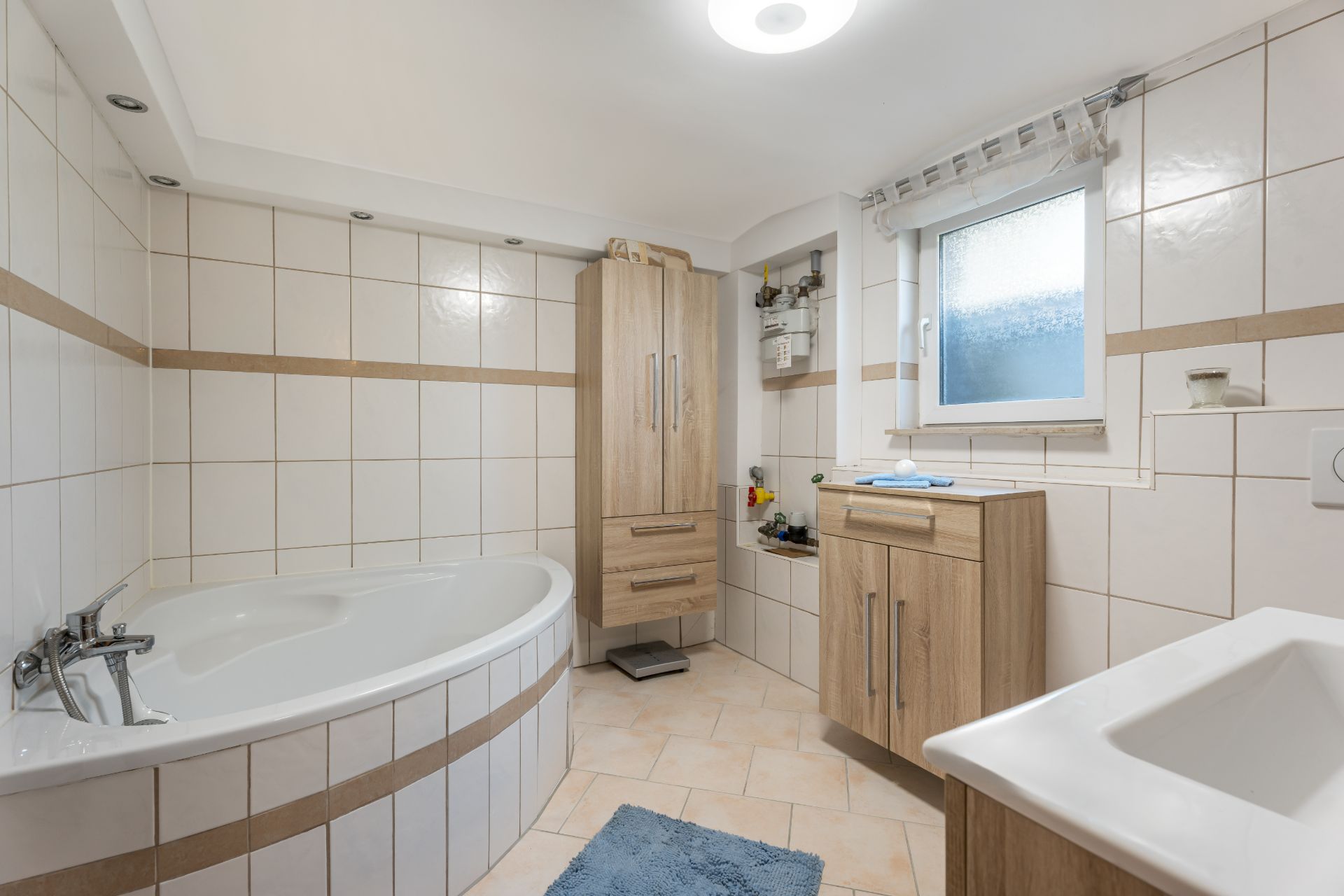 Modernes Badezimmer mit Eckbadewanne, Holzschränken, Fenster und weiß-beigen Fliesen.