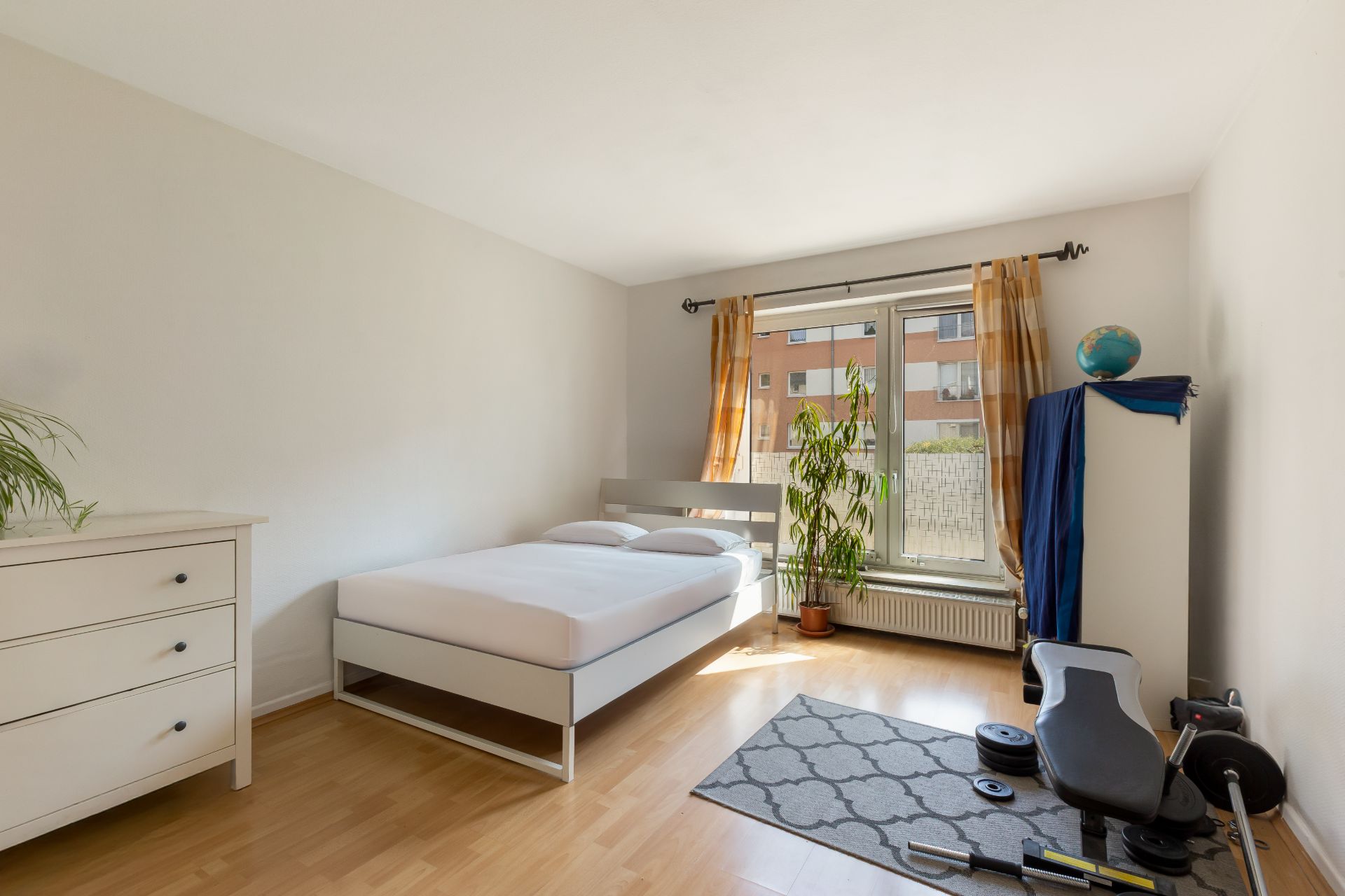 Helles Schlafzimmer mit Bett, Kommode, Schrank, Pflanze und Fitnessgeräten auf Holzboden.