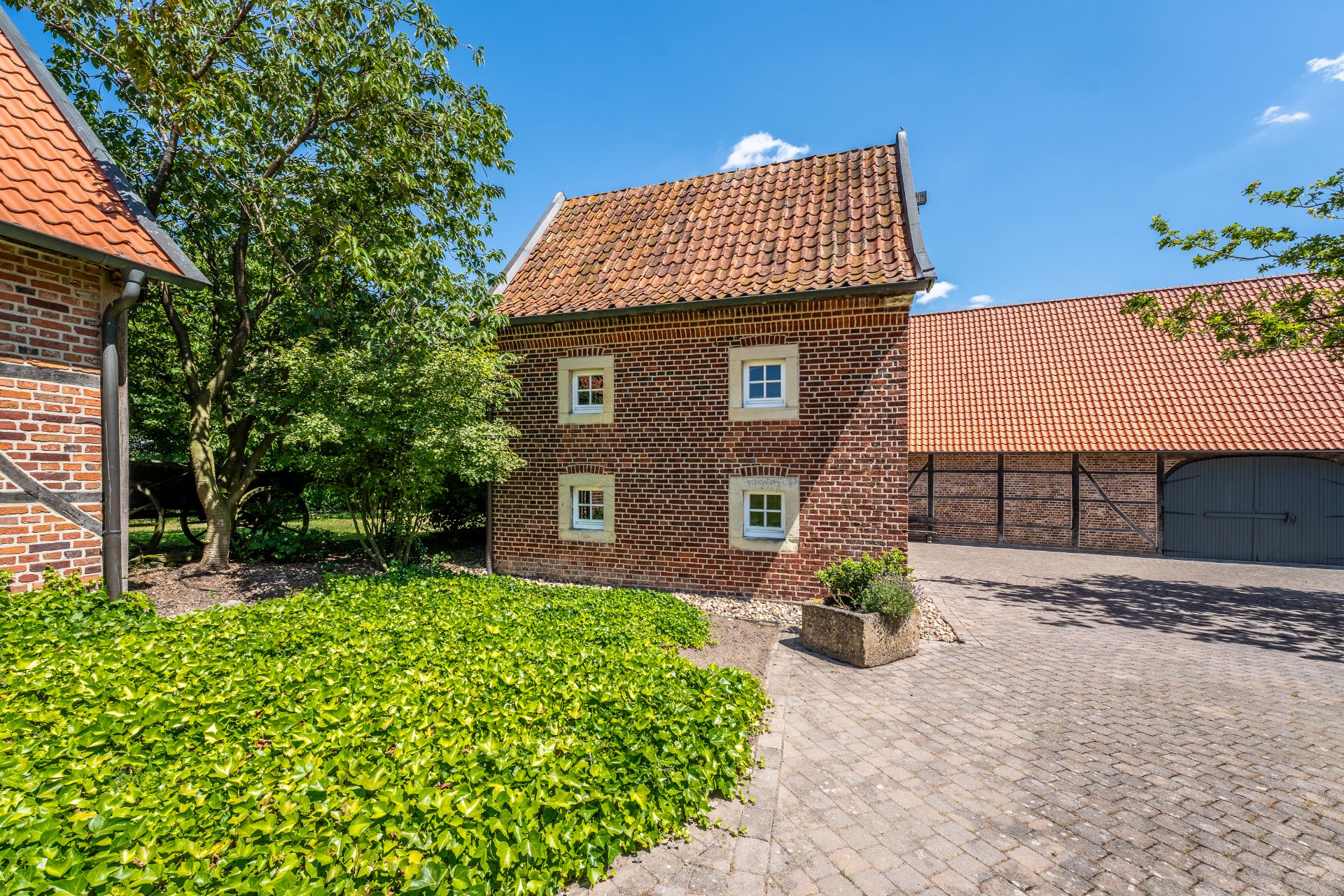Backsteingebäude mit Satteldach, vier Fenstern, gepflastertem Hof und begrüntem Beet im Vordergrund.