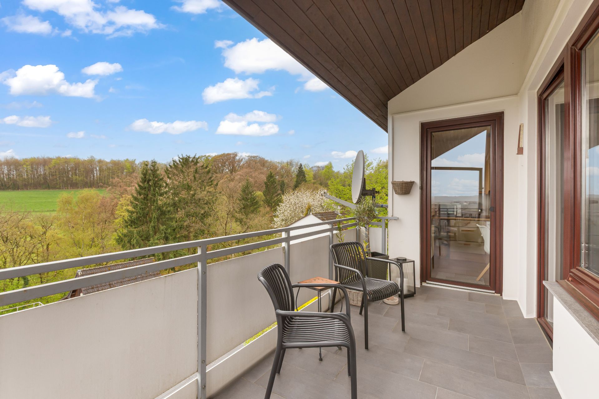 Balkon mit zwei schwarzen Stühlen, Tisch und Blick auf grüne Bäume im Hintergrund.