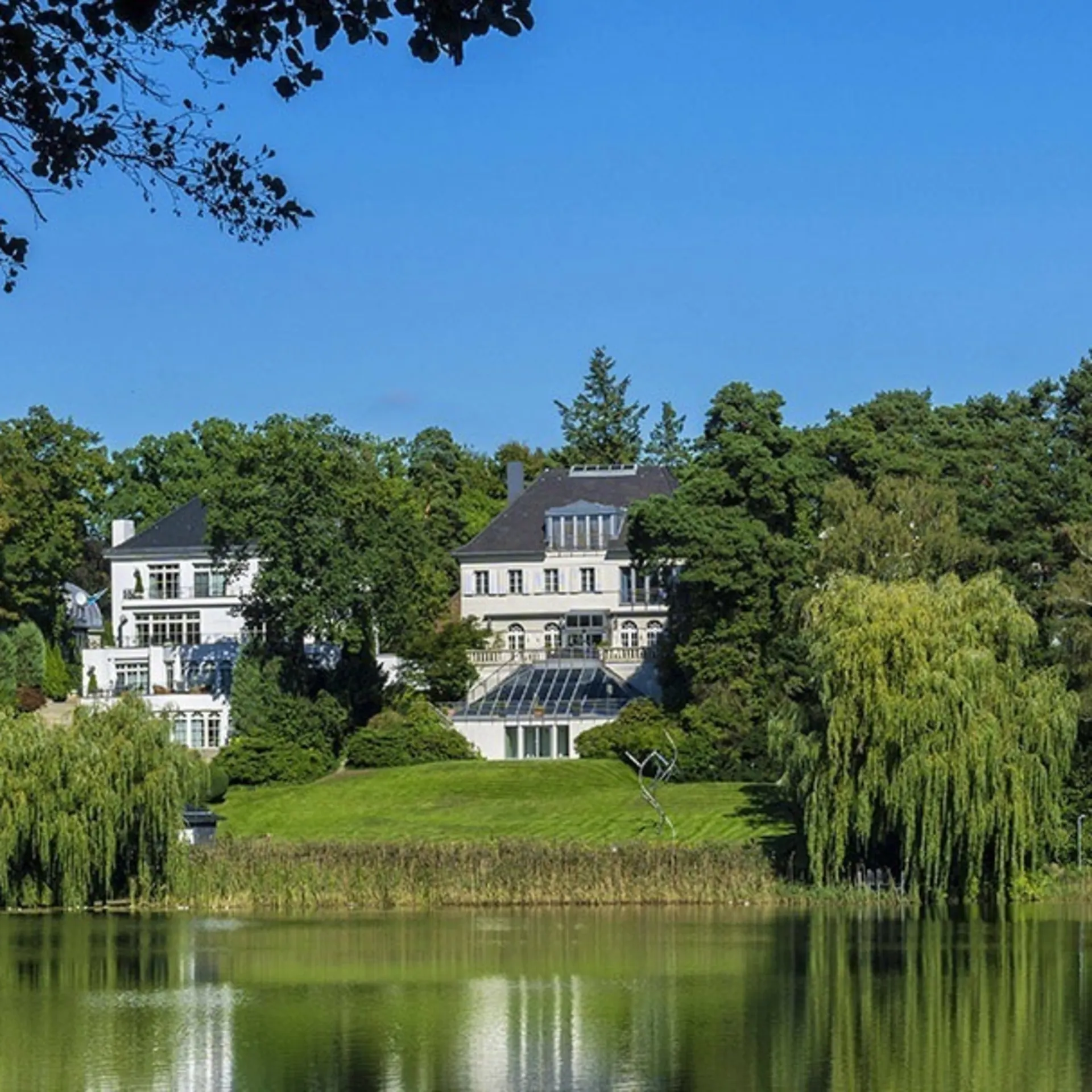 Weiße Villa mit großem Garten am Seeufer, umgeben von Bäumen unter blauem Himmel.