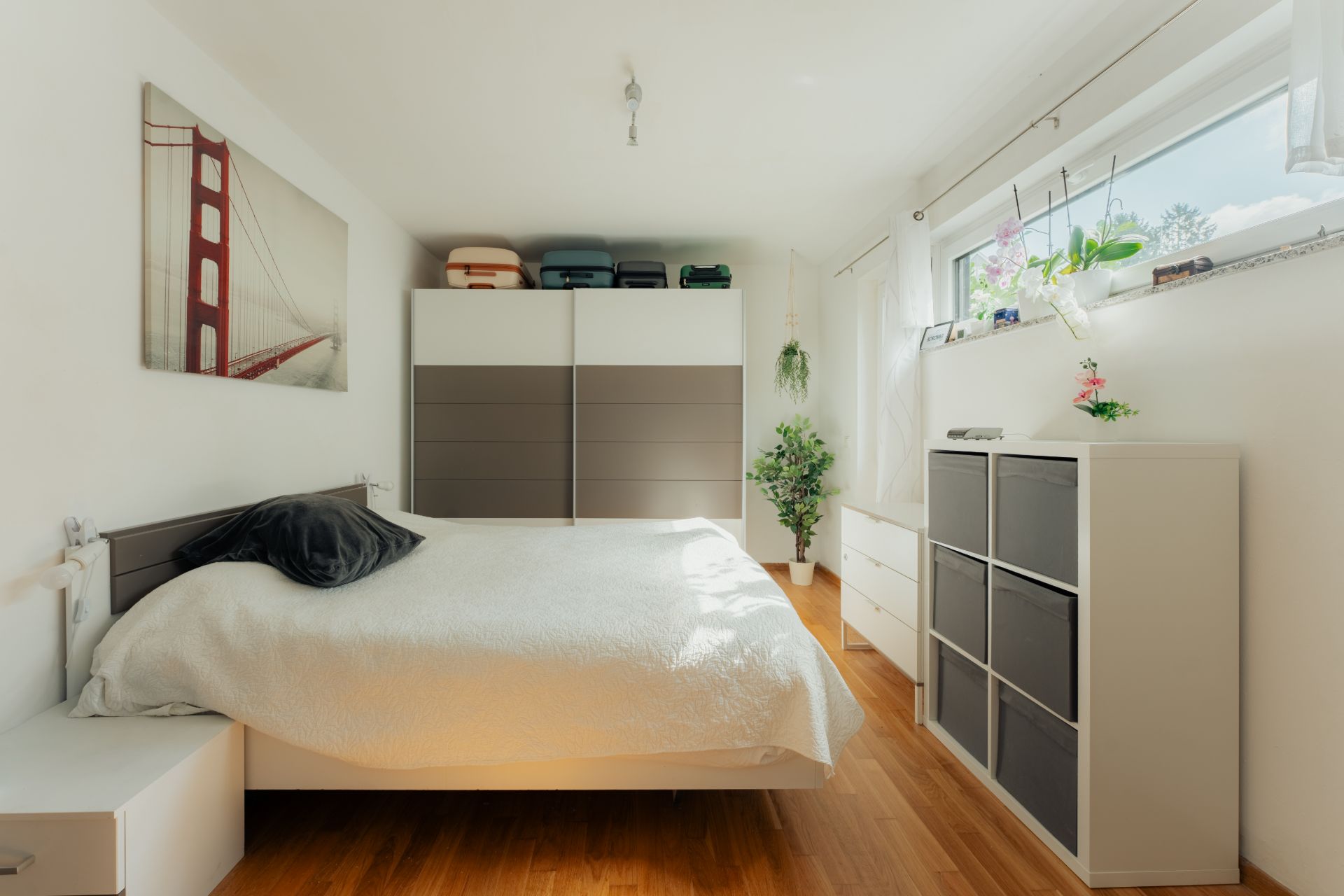 Helles Schlafzimmer mit Doppelbett, Schrank, Kommode und Fenster mit Pflanzen.