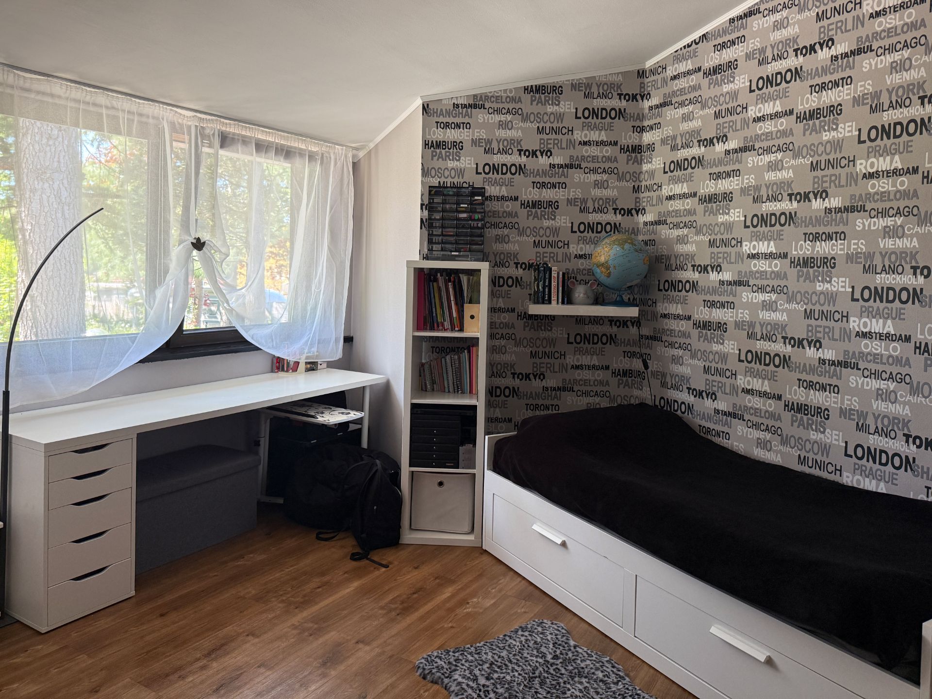 Helles Zimmer mit Bett, Schreibtisch, Regal, Globus und großer Fensterfront mit Vorhängen.