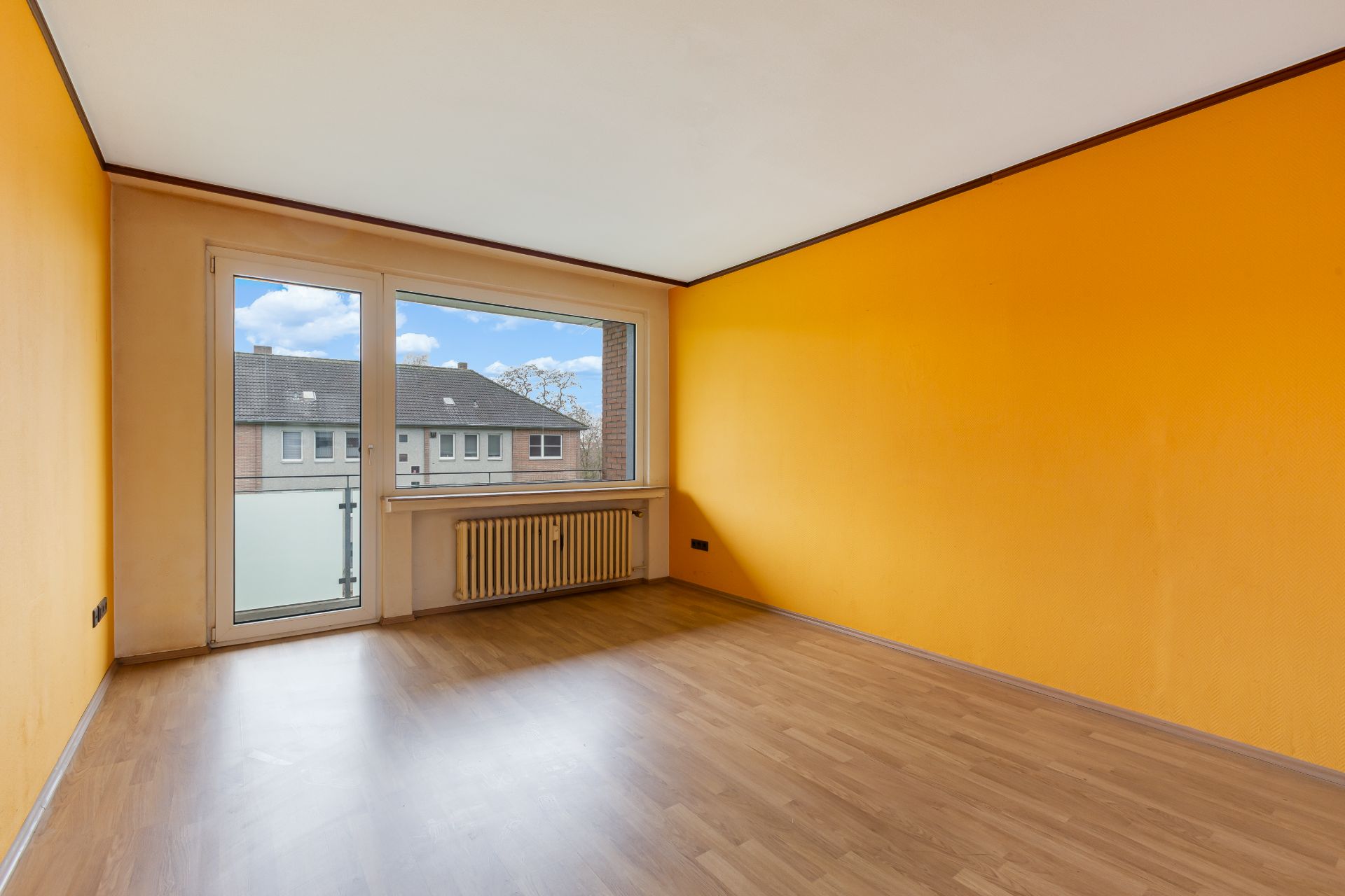 Leerer Raum mit gelben Wänden, Laminatboden und großem Fenster mit Tür zum Balkon.
