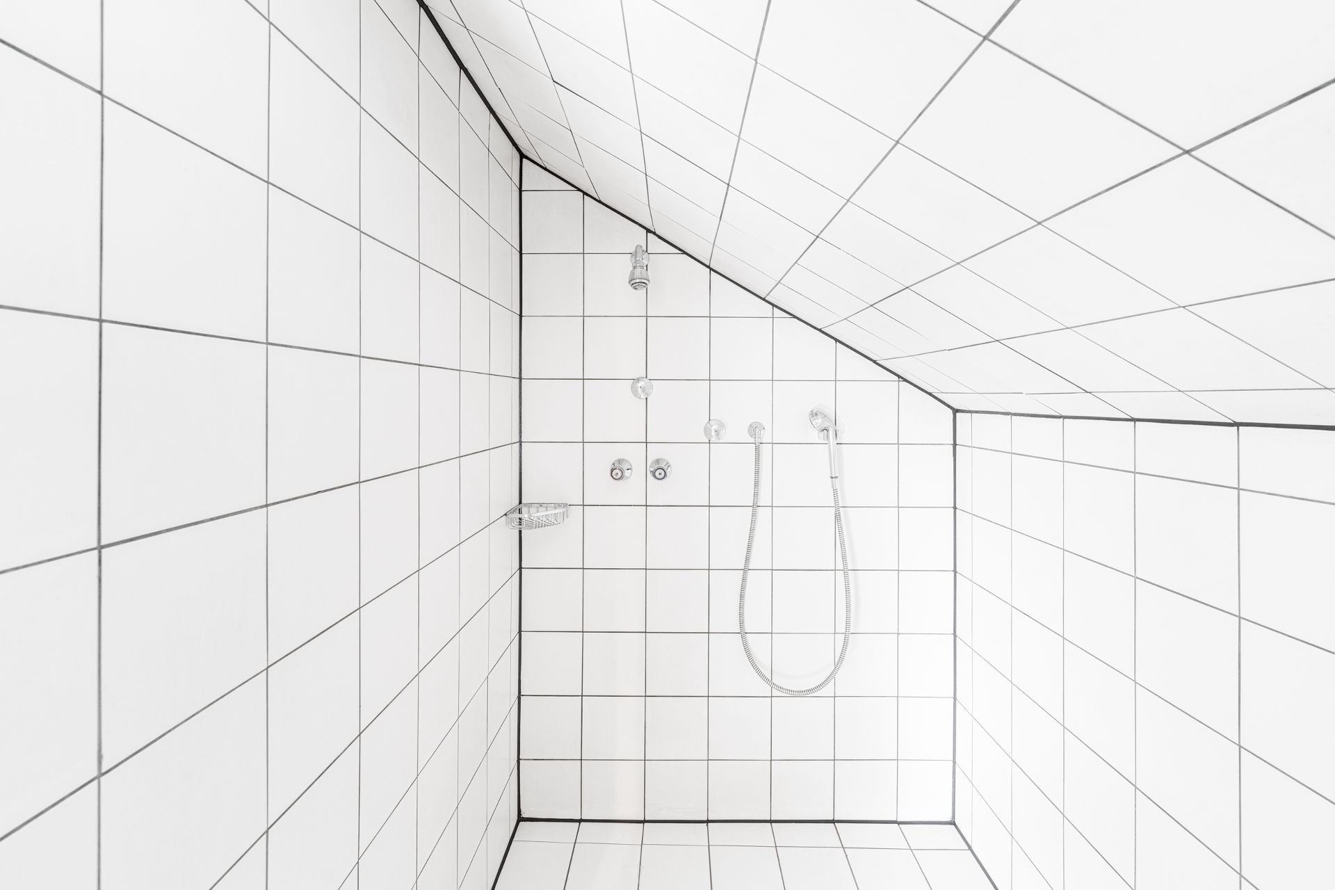Weiße, geflieste Dusche mit schräger Decke und Duschkopf an der Wand.