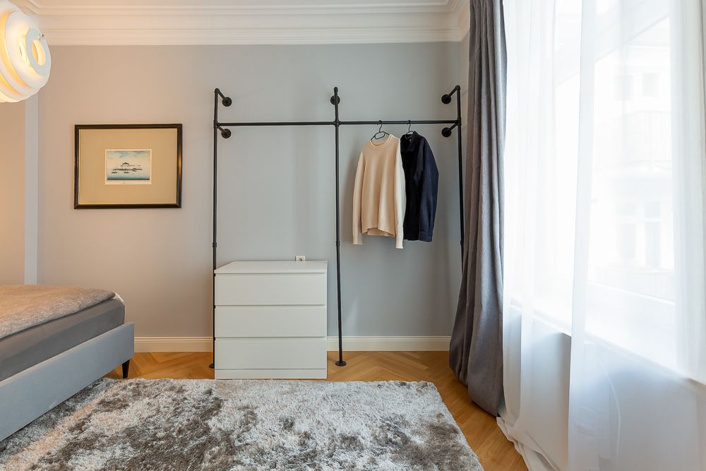 Schlafzimmer mit Kleiderstange, Kommode, Bett und großem Fenster mit Vorhang.