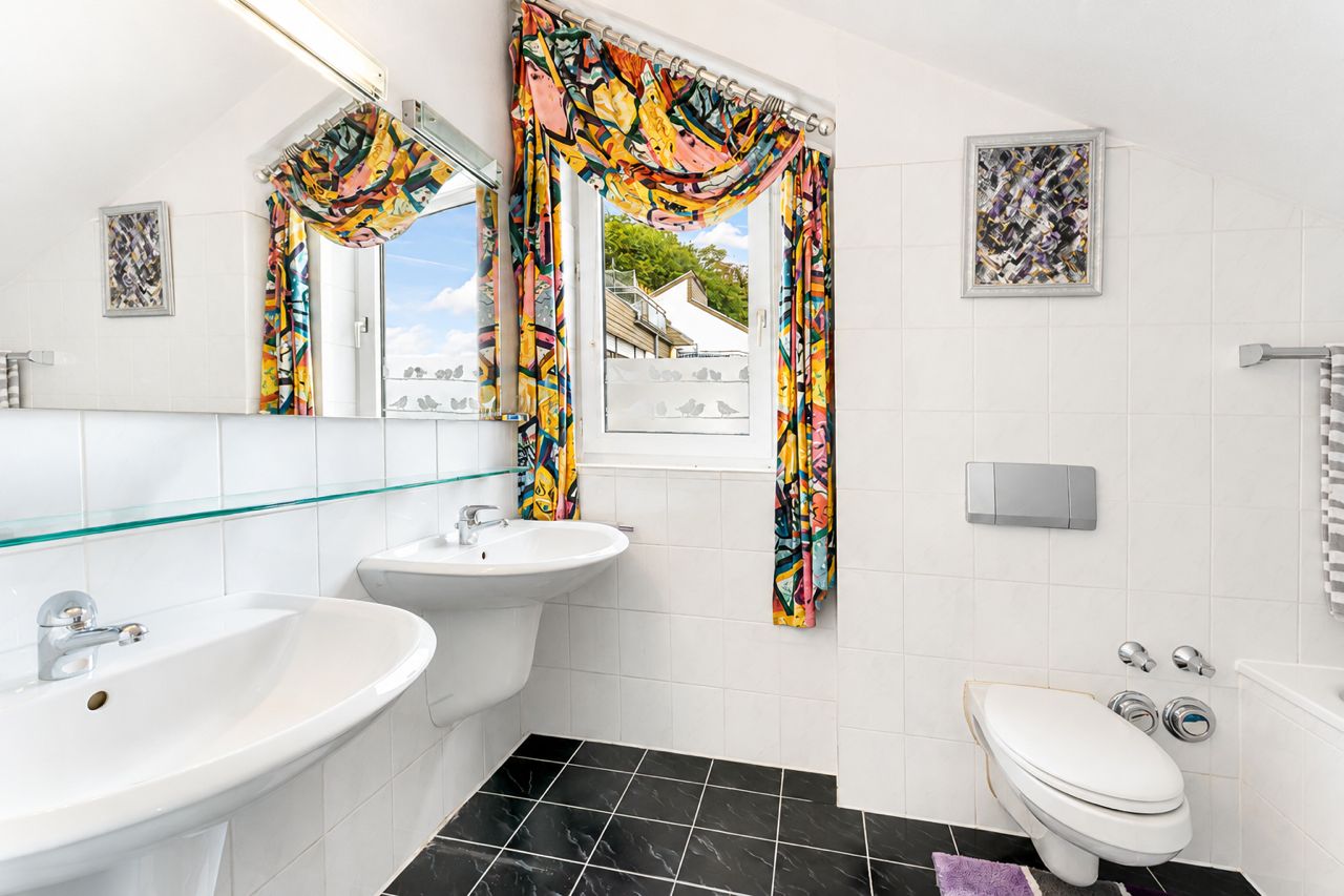 Helles Badezimmer mit zwei Waschbecken, WC, Fenster und bunten Vorhängen.