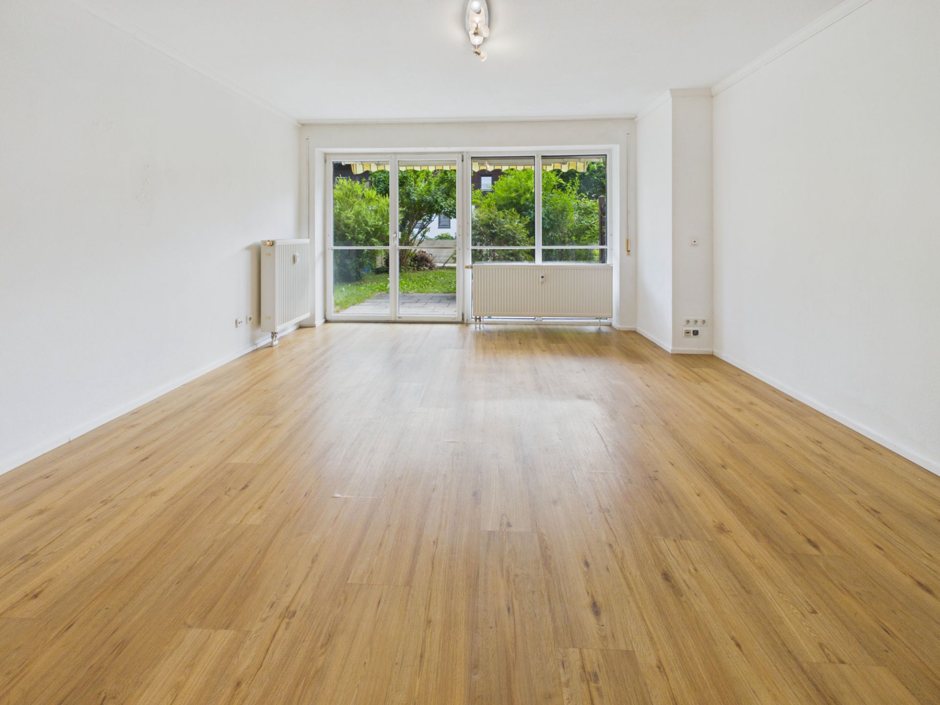Helles, leeres Wohnzimmer mit Holzfußboden und großen Fenstern zum Garten.