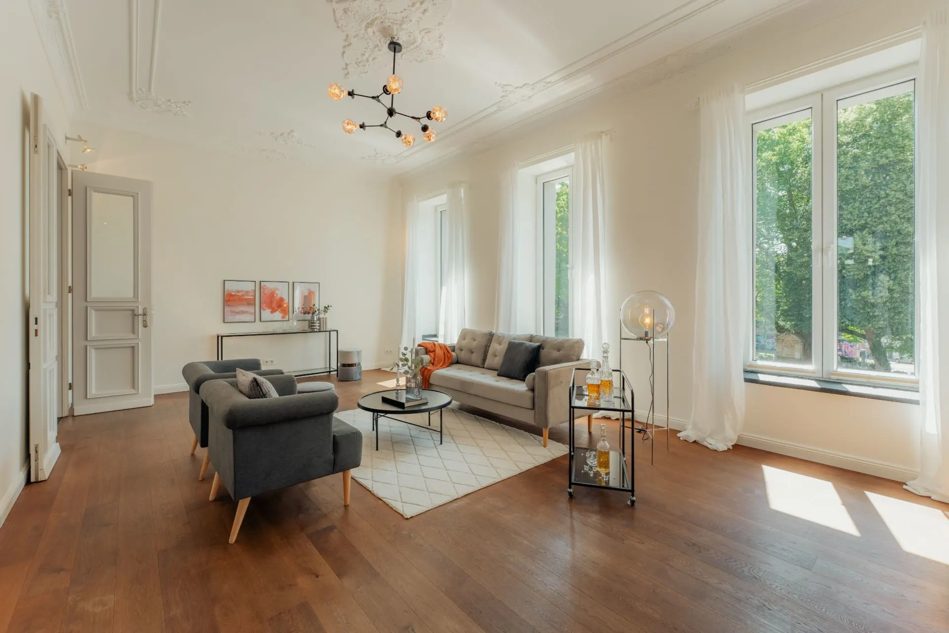 Helles Wohnzimmer mit Holzboden, Sofa, zwei Sesseln und großen Fenstern mit weißen Vorhängen.