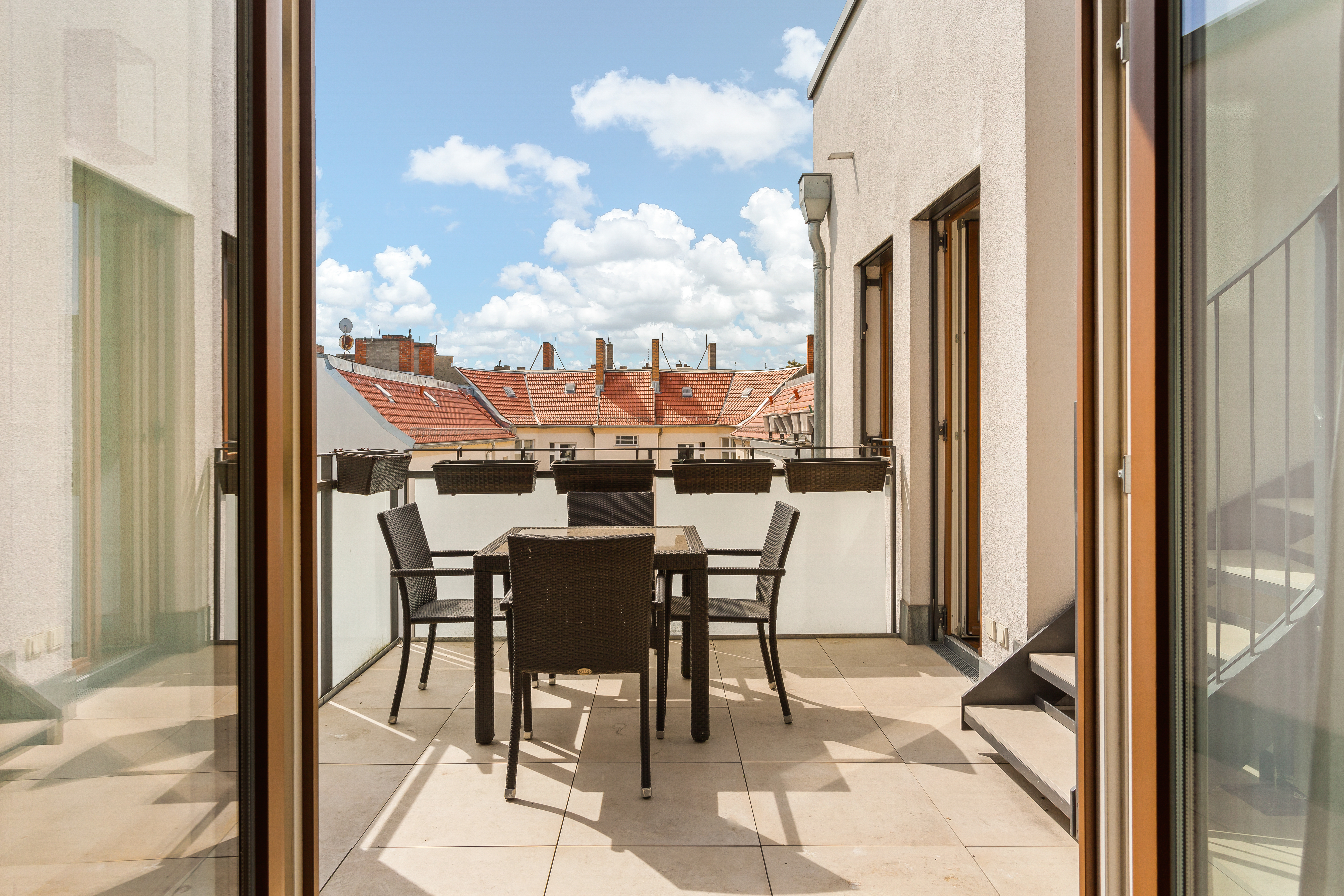 Balkon mit Tisch und vier Stühlen, Blick auf rote Dächer und blauen Himmel mit Wolken.