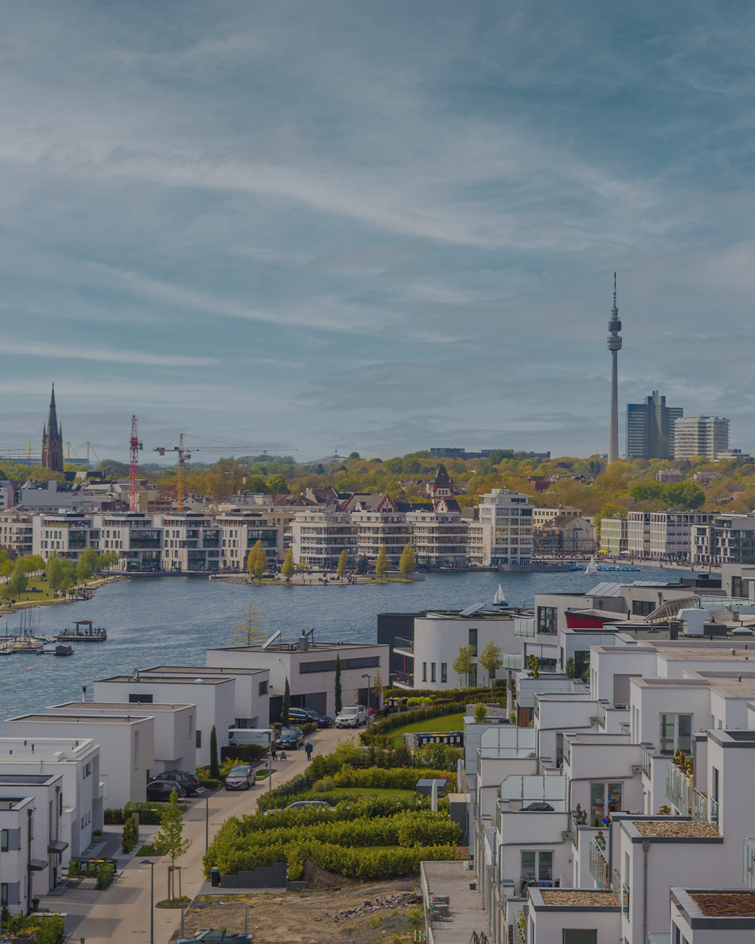 Moderne Wohnhäuser an einem See mit Blick auf Stadt und Fernsehturm im Hintergrund.