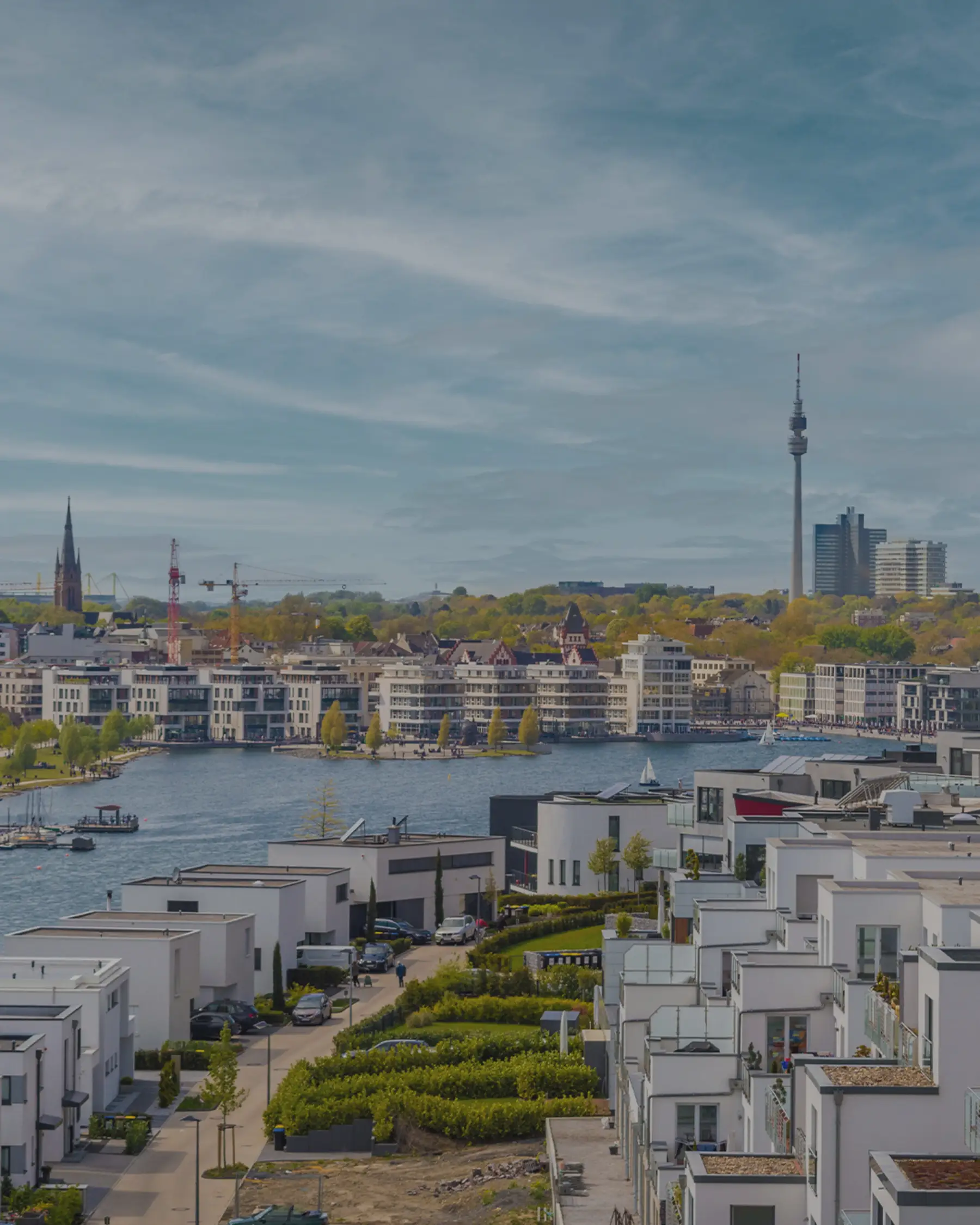 Moderne Wohnhäuser an einem See mit Blick auf Stadt und Fernsehturm im Hintergrund.