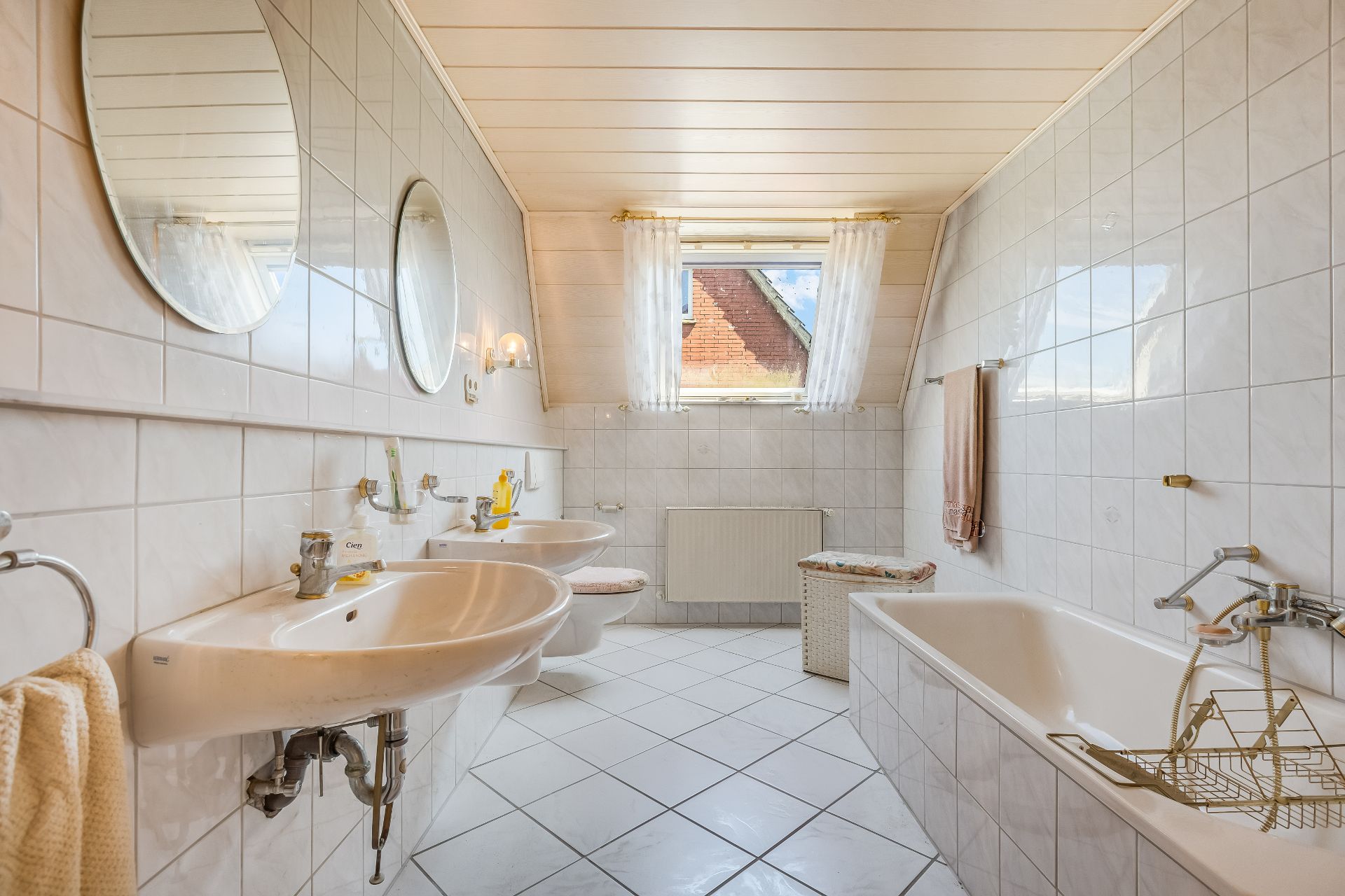 Helles Bad mit zwei Waschbecken, Badewanne, zwei Spiegeln und Fenster mit Vorhängen.