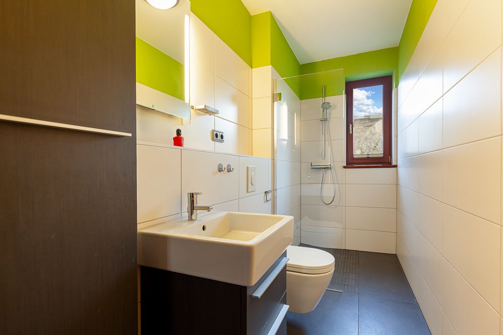 Modernes Bad mit Waschbecken, Toilette, Dusche und Fenster, weiße Fliesen und grüne Wände.