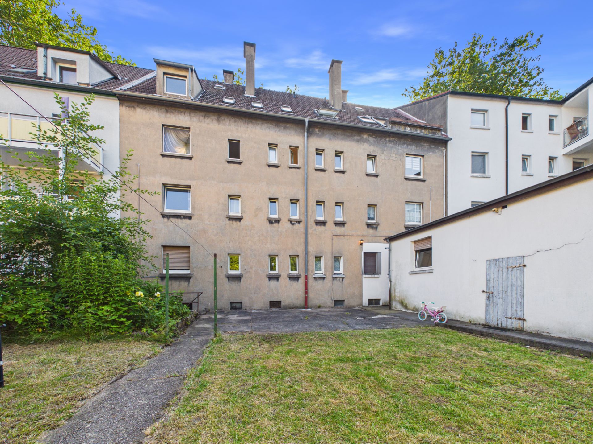 Hinterhof eines Mehrfamilienhauses mit Rasenfläche, Weg und einem kleinen Schuppen.