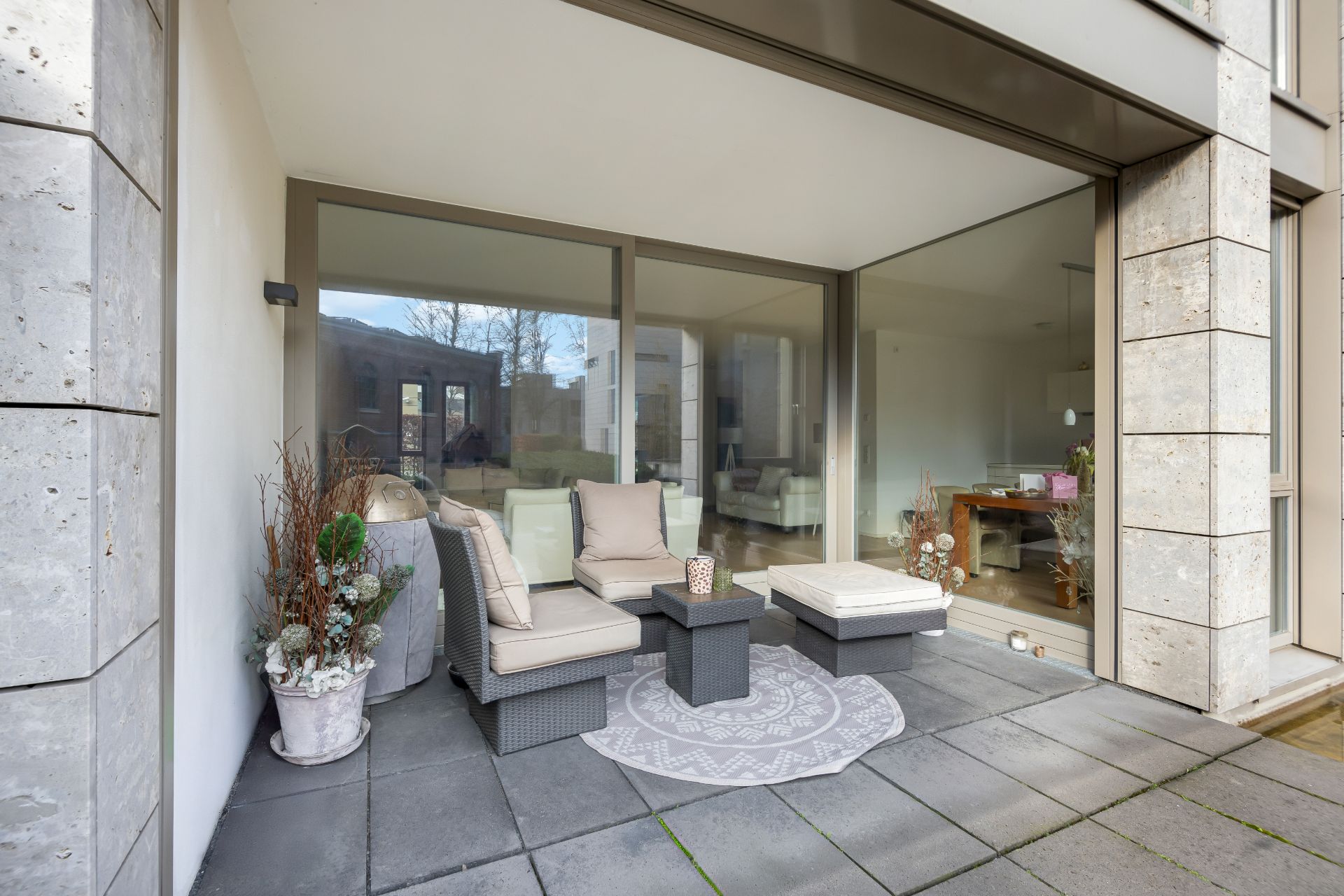 Überdachte Terrasse mit Sitzgruppe, Pflanzen und Blick ins Wohnzimmer durch große Glasfront.