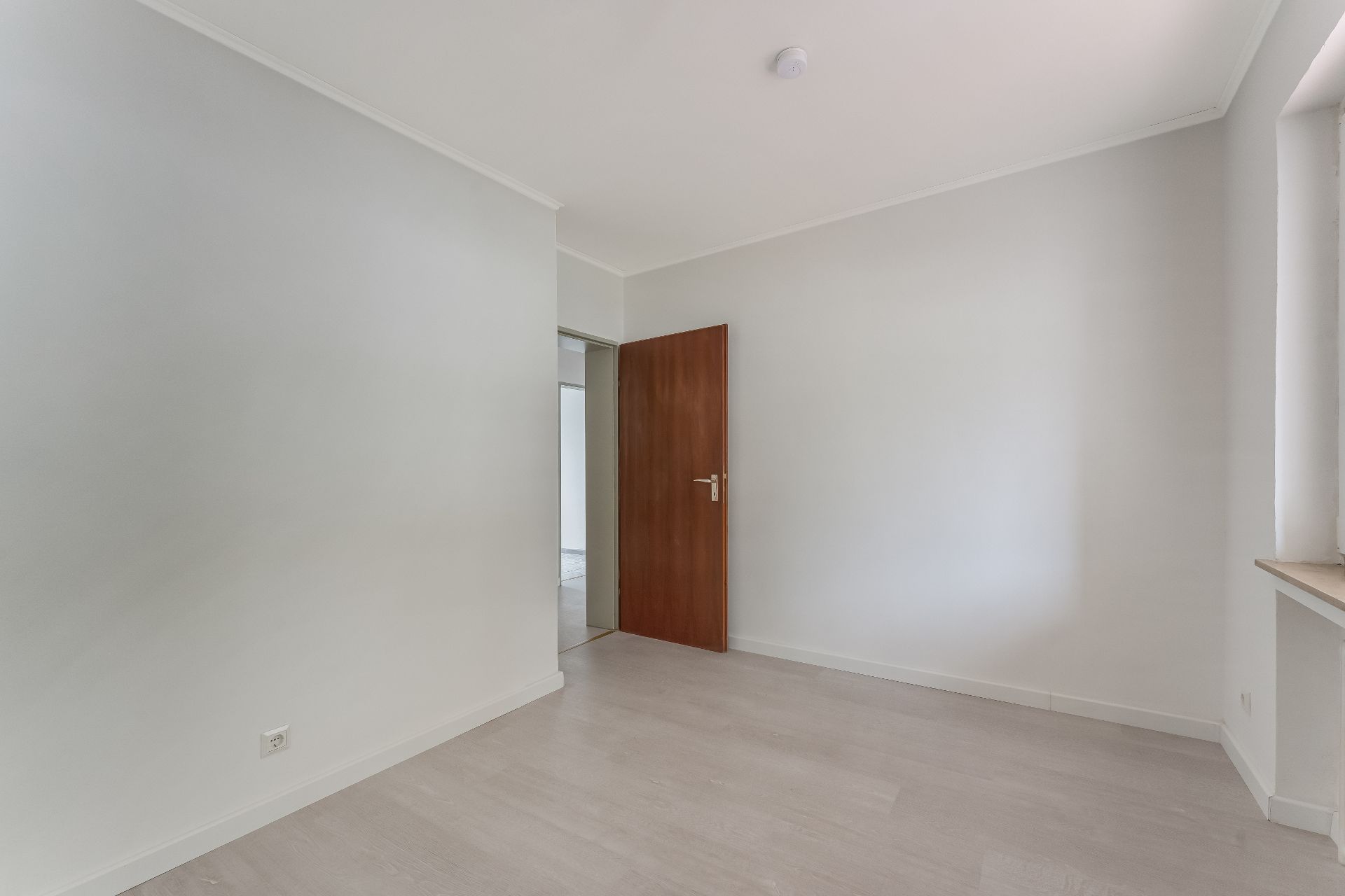 Leeres Zimmer mit weiß gestrichenen Wänden, Holzfußboden, einem Fenster und einer braunen Tür.