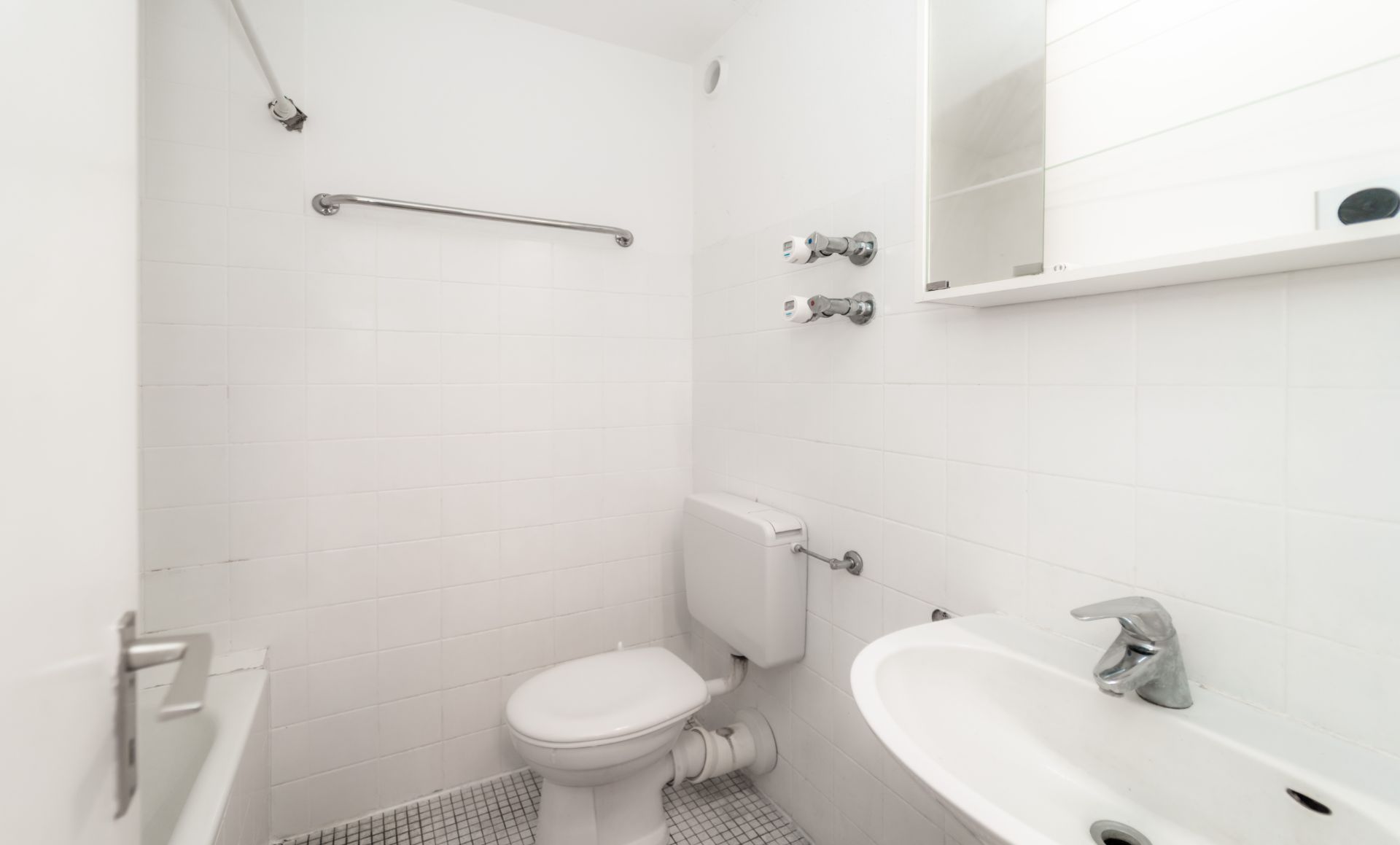 Kleines, weiß gefliestes Badezimmer mit Toilette, Waschbecken und Spiegel.