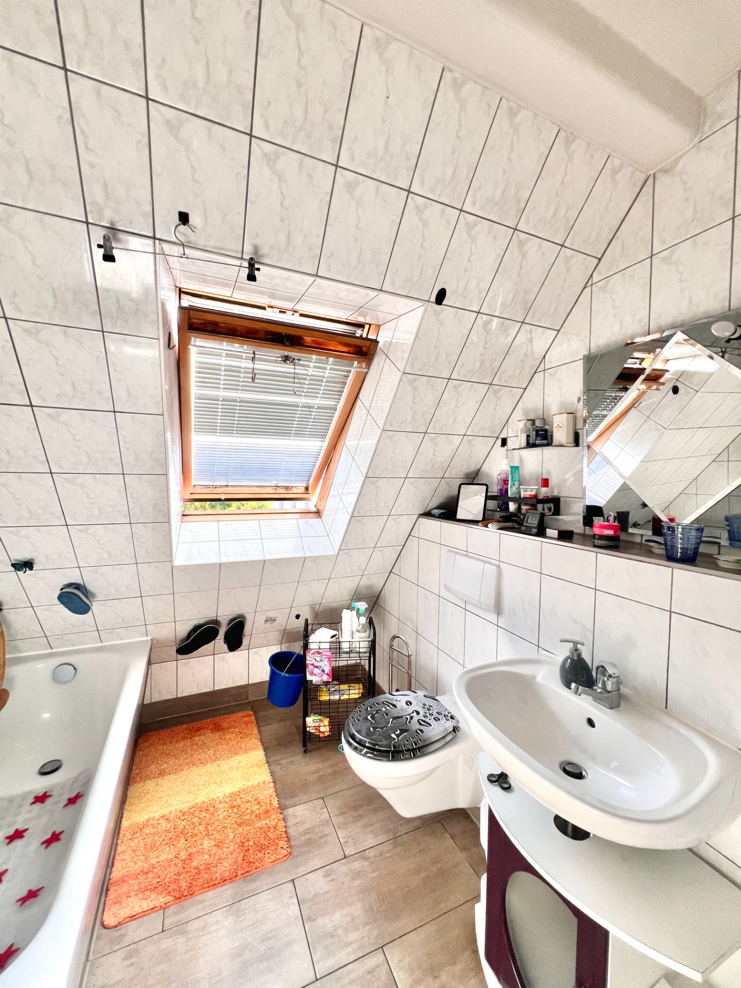 Helles Badezimmer mit Dachfenster, Badewanne, Waschbecken, Toilette und Regalen mit Pflegeartikeln.