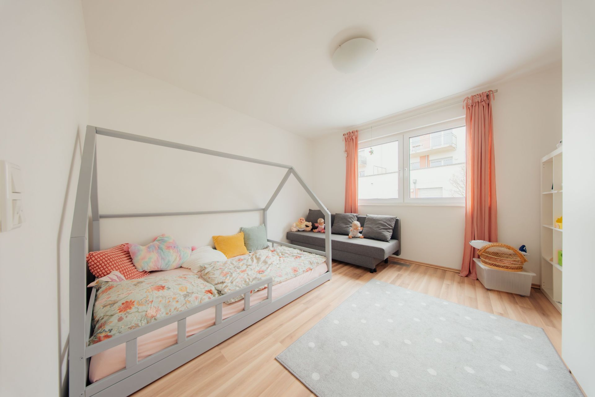 Helles Kinderzimmer mit Hausbett, Sofa, Teppich, Regalen und Fenstern mit rosa Vorhängen.