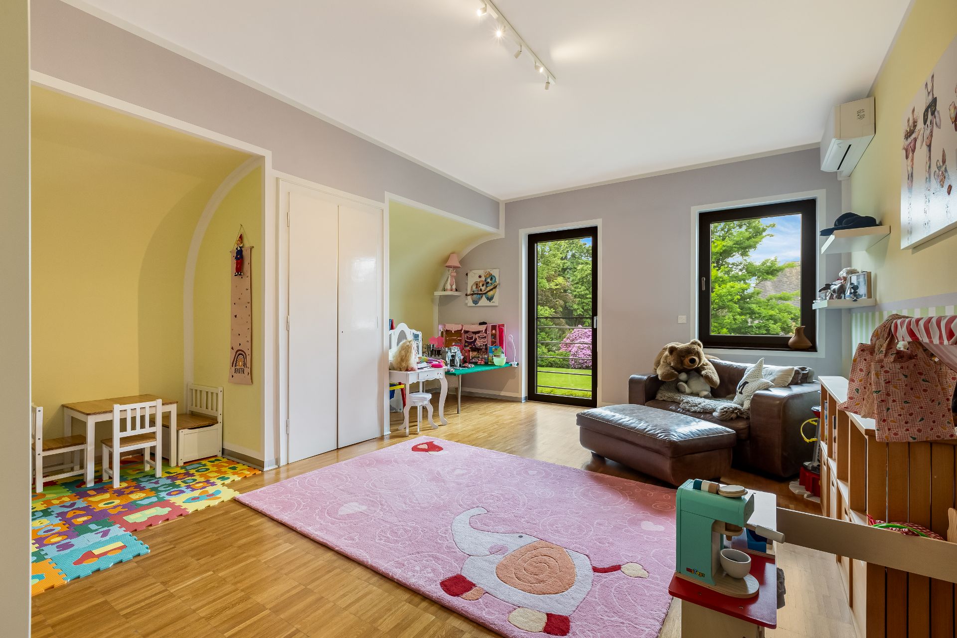 Helles Kinderzimmer mit Spielzeug, kleinem Tisch, Sofa und großem Fenster mit Gartenblick.