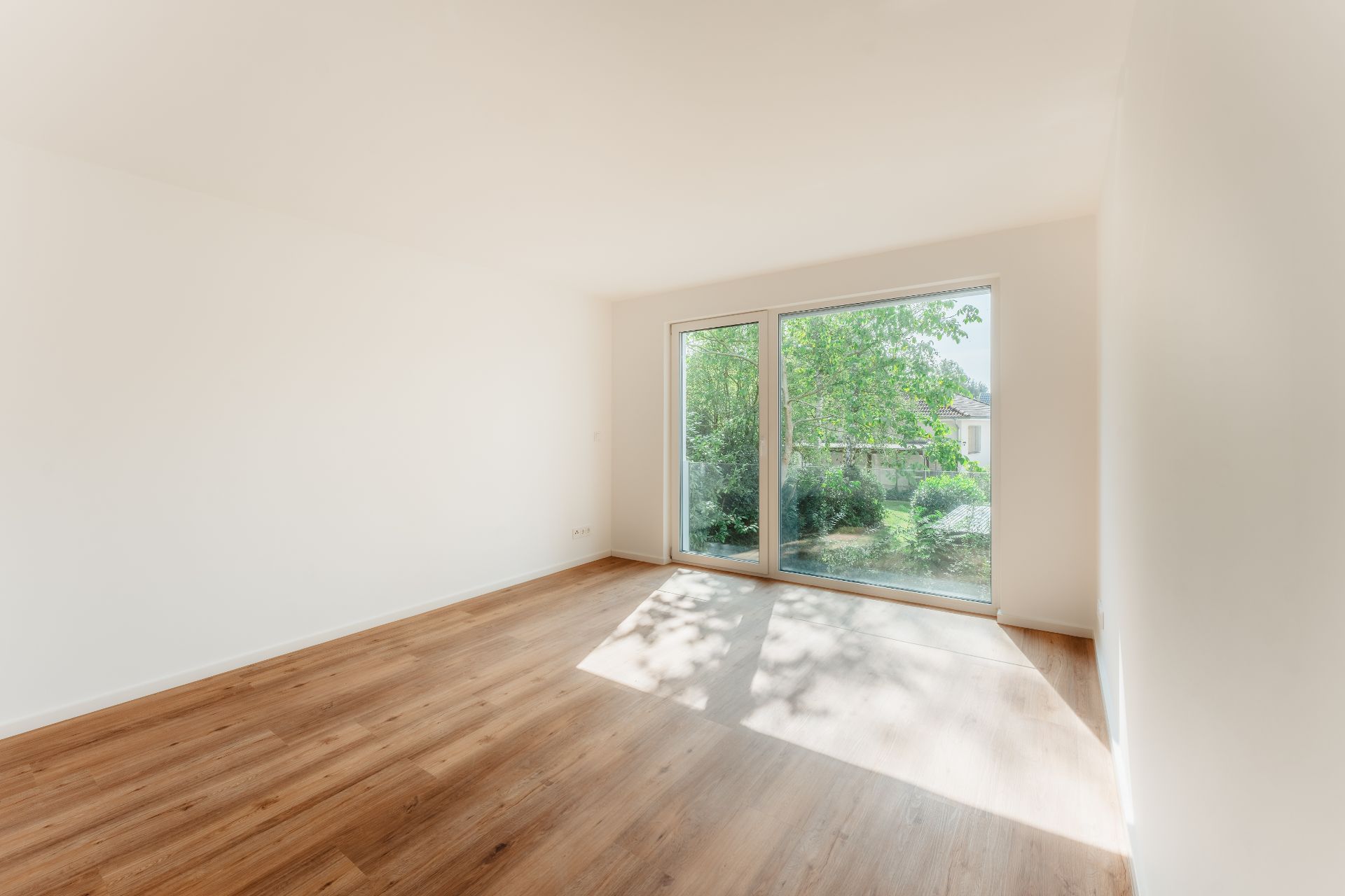 Helles, leeres Zimmer mit Holzfußboden und großen Fenstern zum Garten.