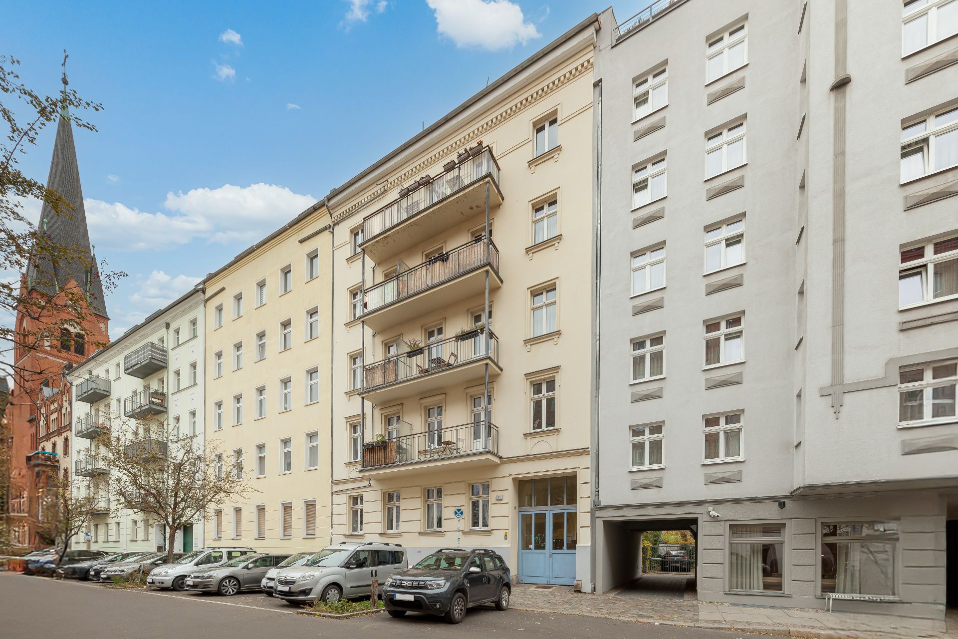 Mehrstöckiges Wohnhaus mit Balkonen und parkenden Autos davor, daneben eine Kirche mit Turm.