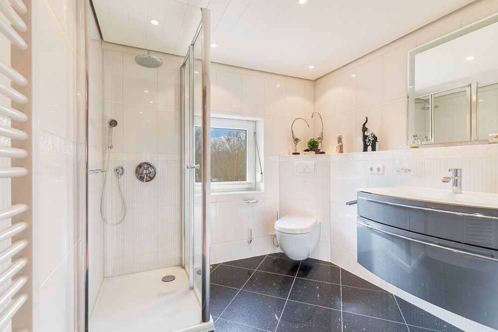 Modernes Bad mit Dusche, WC, Waschbecken und Spiegel, schwarze Fliesen, Fenster an der Wand.