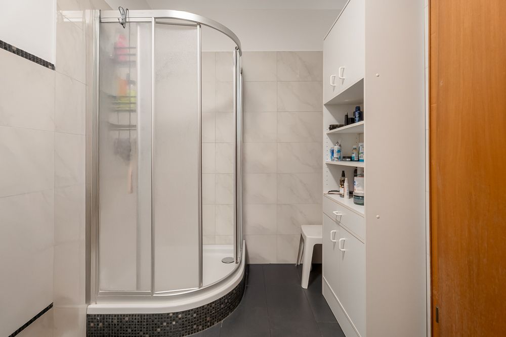 Badezimmer mit Eckdusche, weißen Fliesen und offenen Regalen mit Pflegeartikeln.