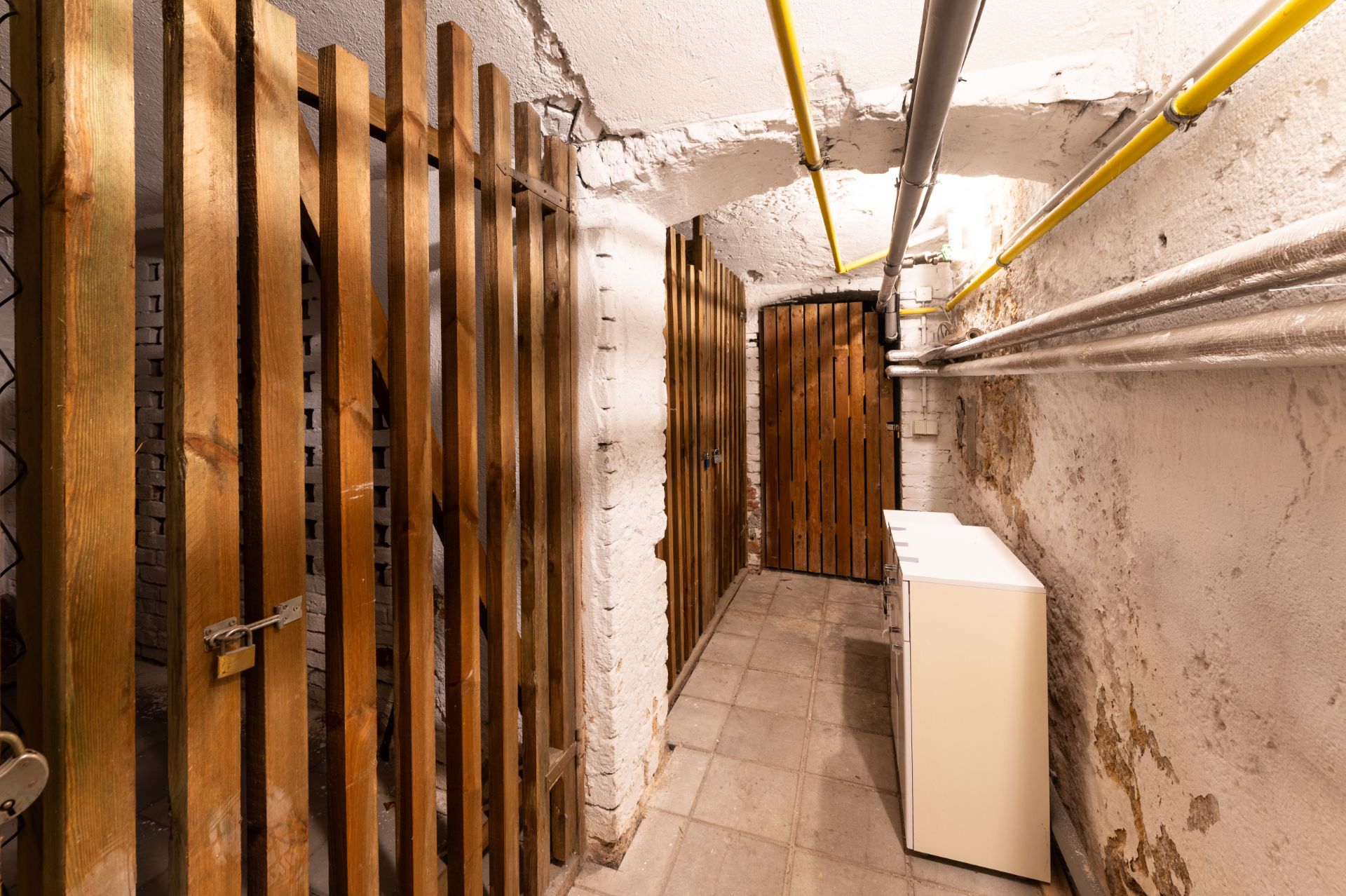 Ein Kellerraum mit Holzgittertüren, weißen Wänden, Rohren an der Decke und einem kleinen weißen Schrank.