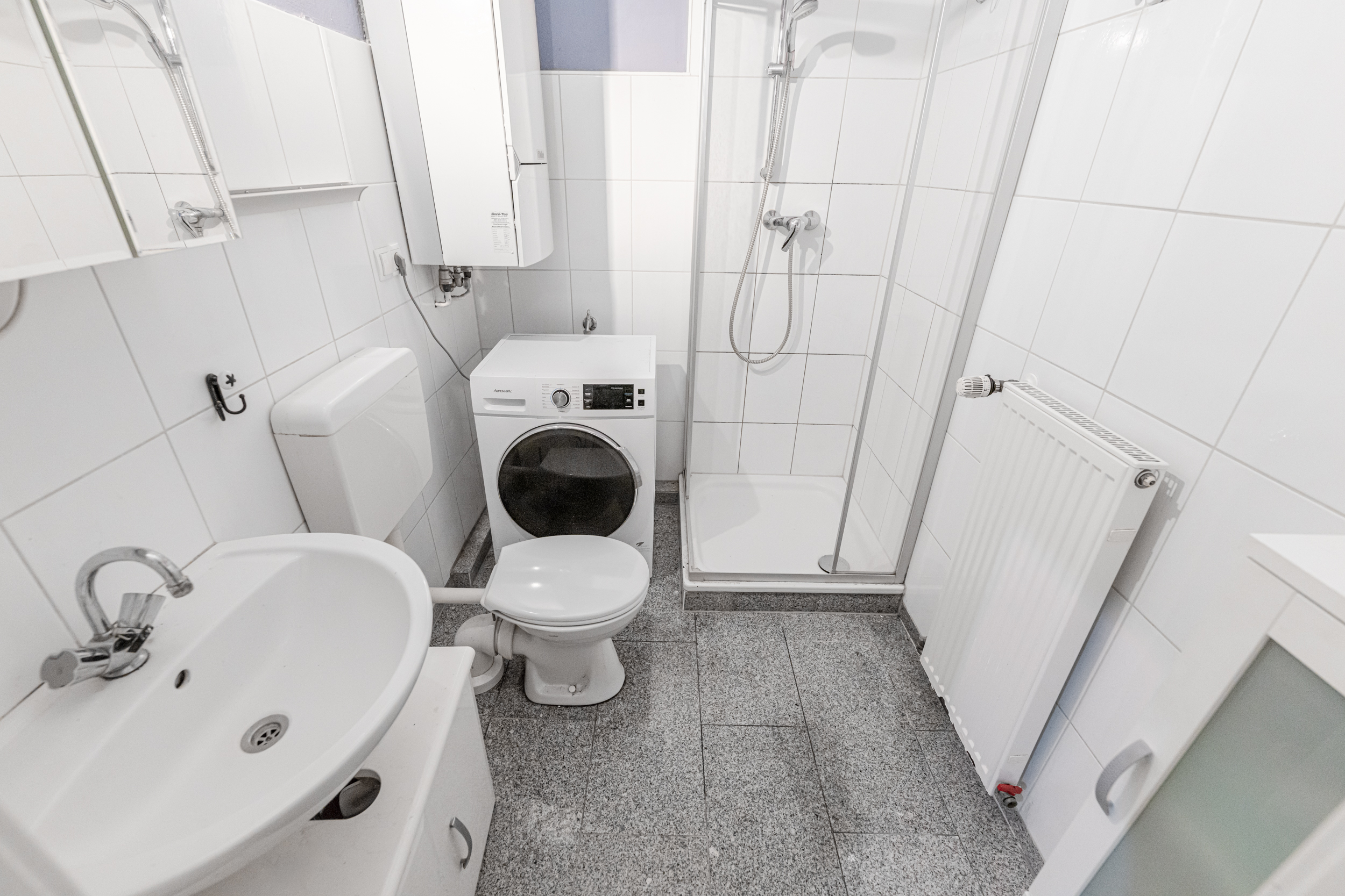 Kleines Badezimmer mit Waschbecken, Toilette, Dusche, Waschmaschine und Heizkörper.