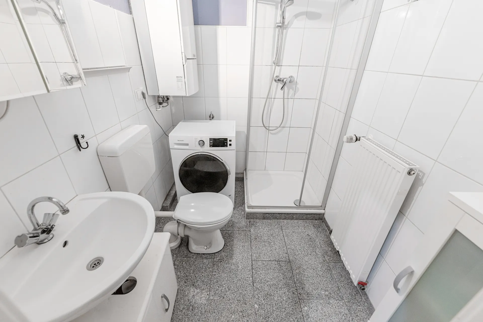 Kleines Badezimmer mit Waschbecken, Toilette, Dusche, Waschmaschine und Heizkörper.