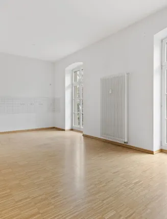 Heller leerer Raum mit Parkettboden, weißen Wänden und großen Fenstern mit Blick nach draußen.