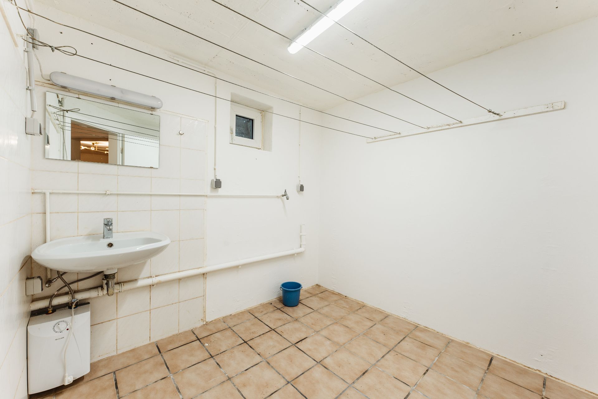 Heller Raum mit Waschbecken, Spiegel, Fliesenboden und Wäscheleinen an der Decke.