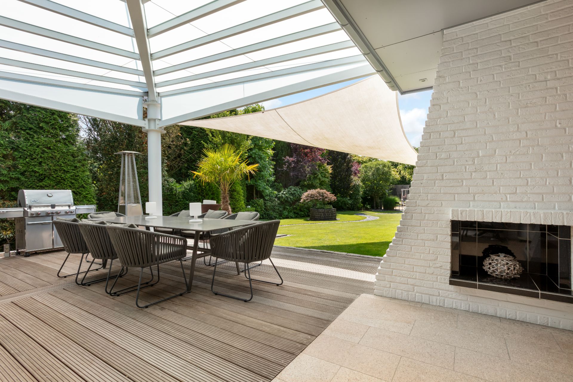 Überdachte Terrasse mit Esstisch, Stühlen und Grill, Blick auf einen Garten mit Rasen.