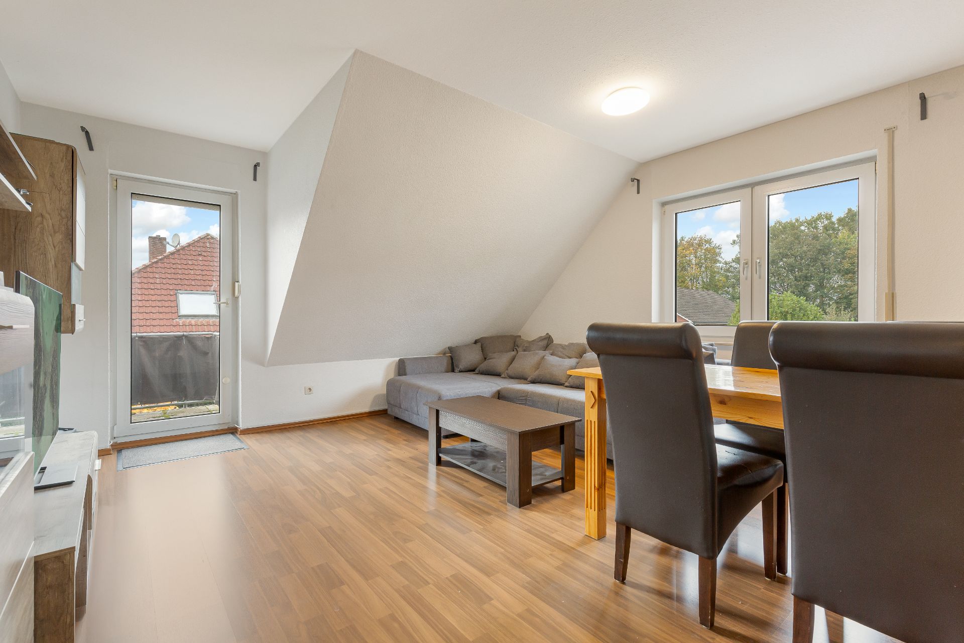 Wohnzimmer mit Holzfußboden, grauem Sofa, Esstisch und Zugang zum Balkon.