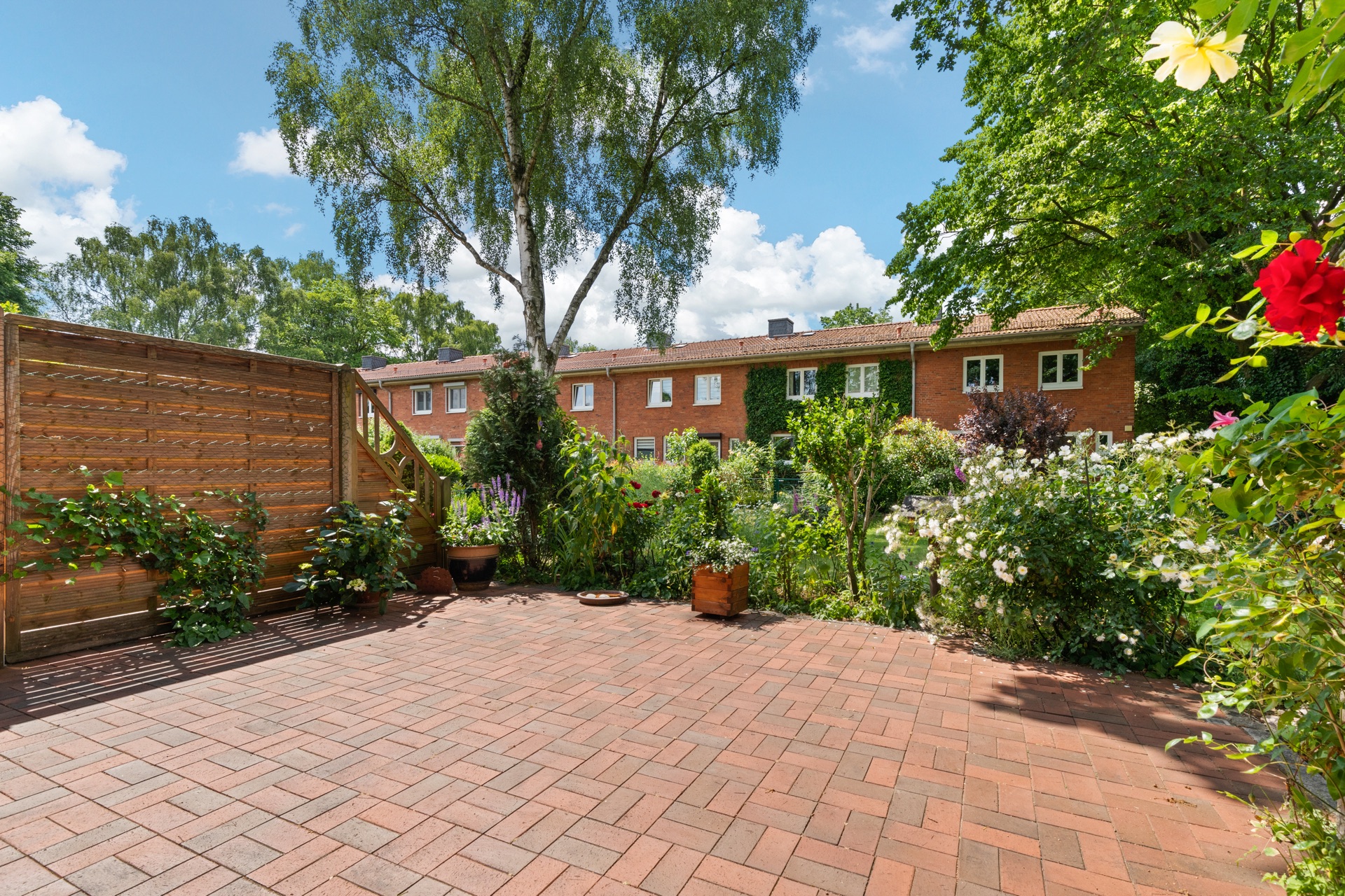 Gepflasterte Terrasse mit Holzsichtschutz, Pflanzen und Blick auf einen grünen Garten.