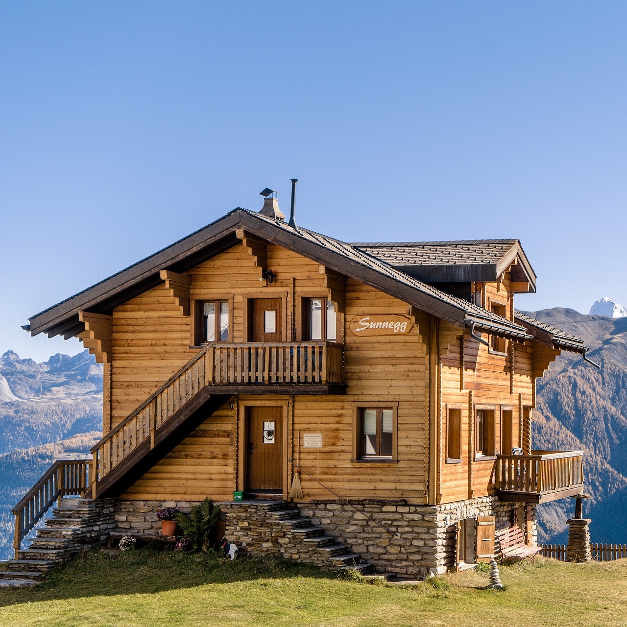 Zweistöckiges Holzhaus mit Steintreppe und Balkon in bergiger Landschaft.