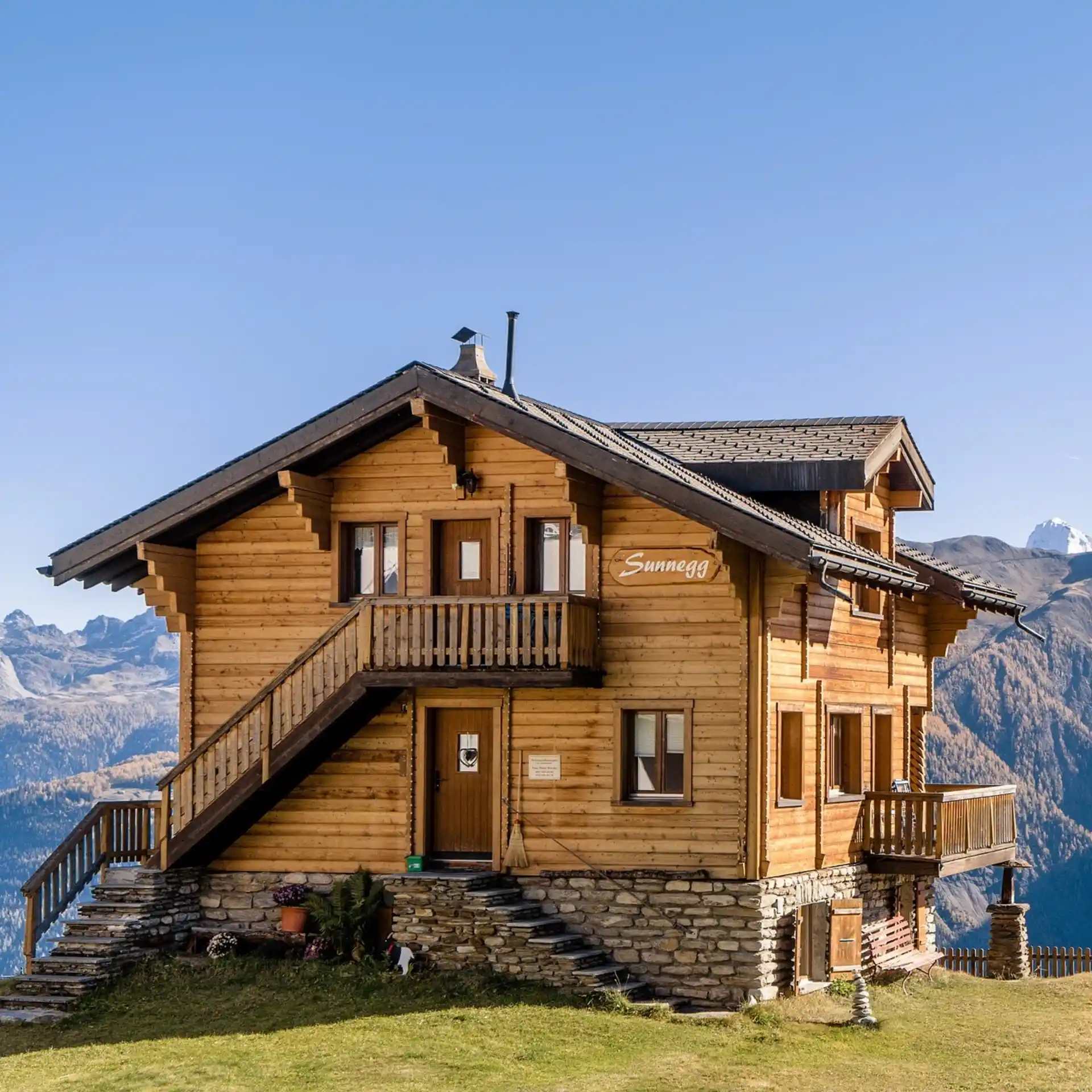 Zweistöckiges Holzhaus mit Steintreppe und Balkon in bergiger Landschaft.