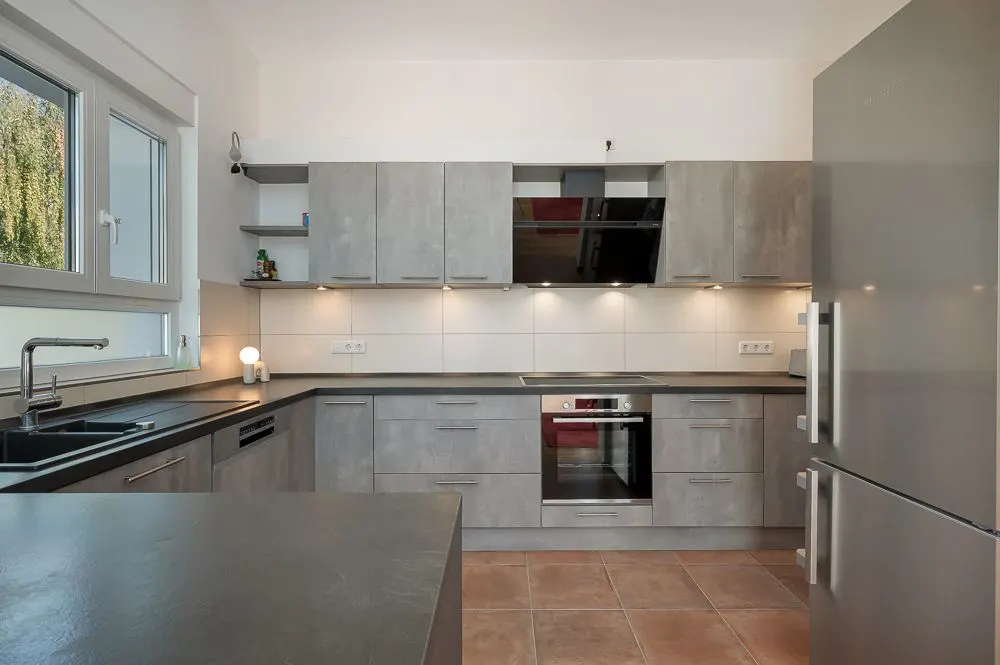 Moderne Einbauküche mit grauen Schränken, großem Kühlschrank und Fenster auf der linken Seite.